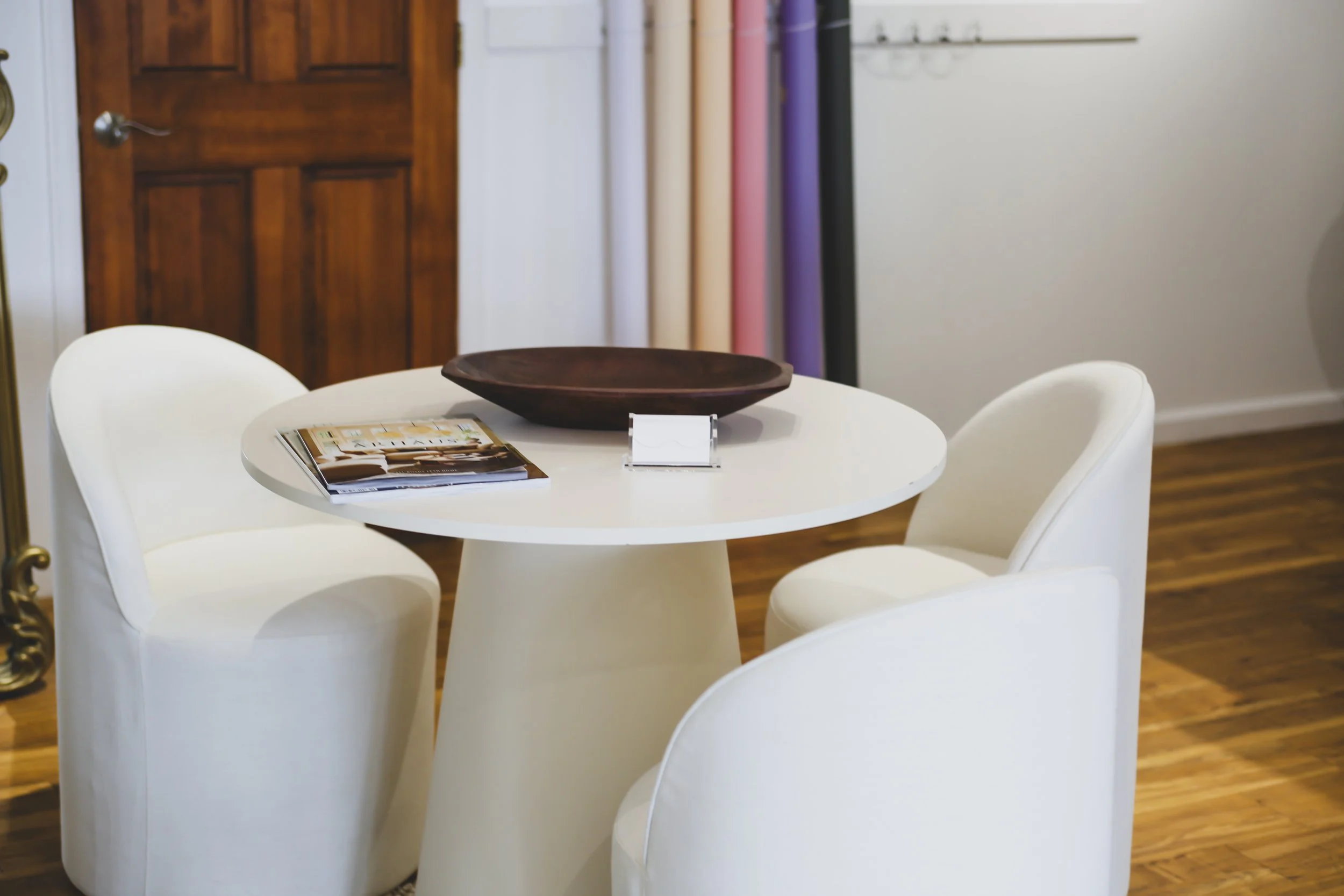 A white round table surrounded by four white chairs in a room with wooden flooring and a door in the background. On the table, there is a dark wooden bowl, a magazine, and a small stand with a white card.