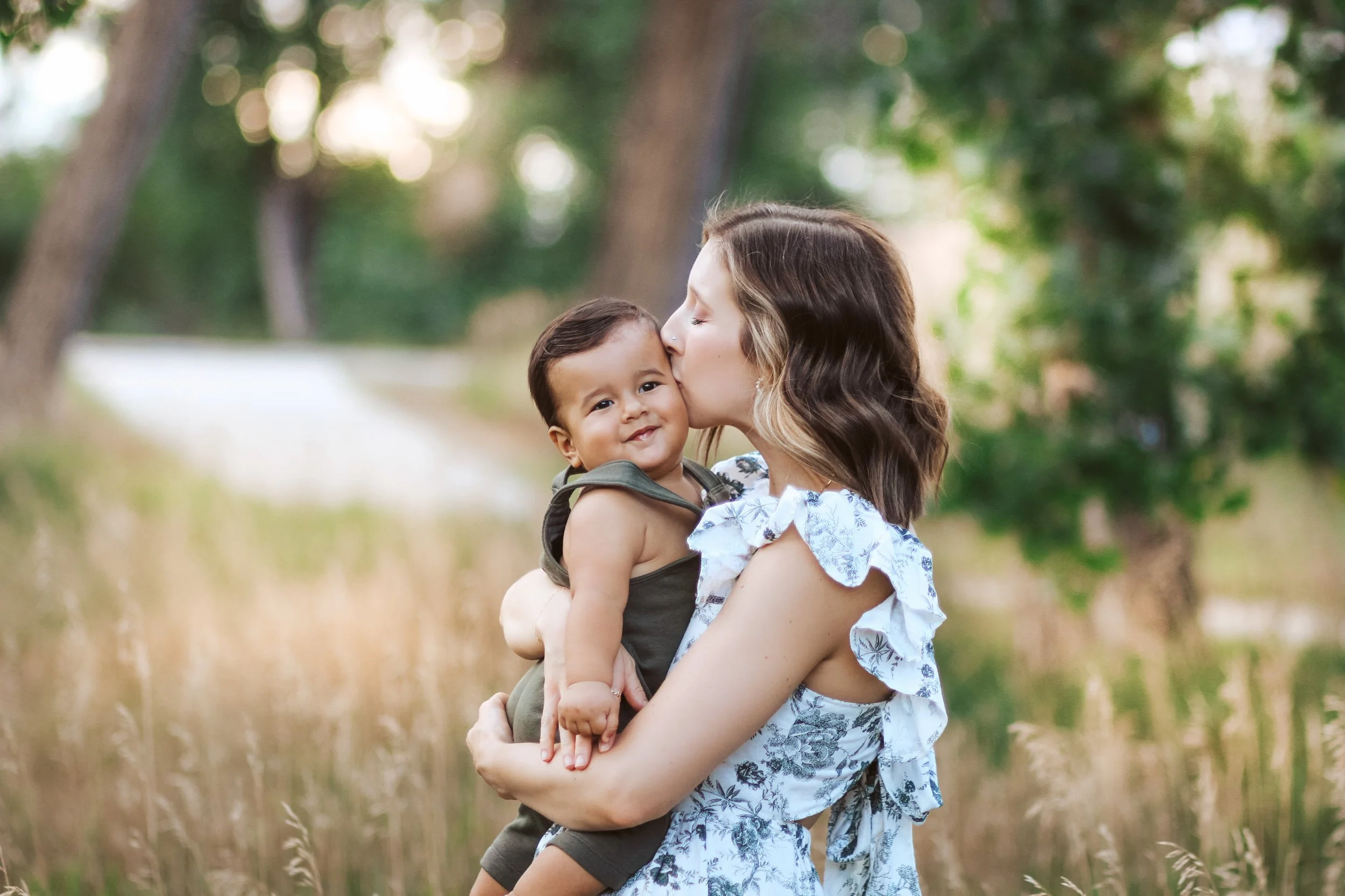 A woman kissing a smiling young child on the cheek outdoors in a field with trees in the background.