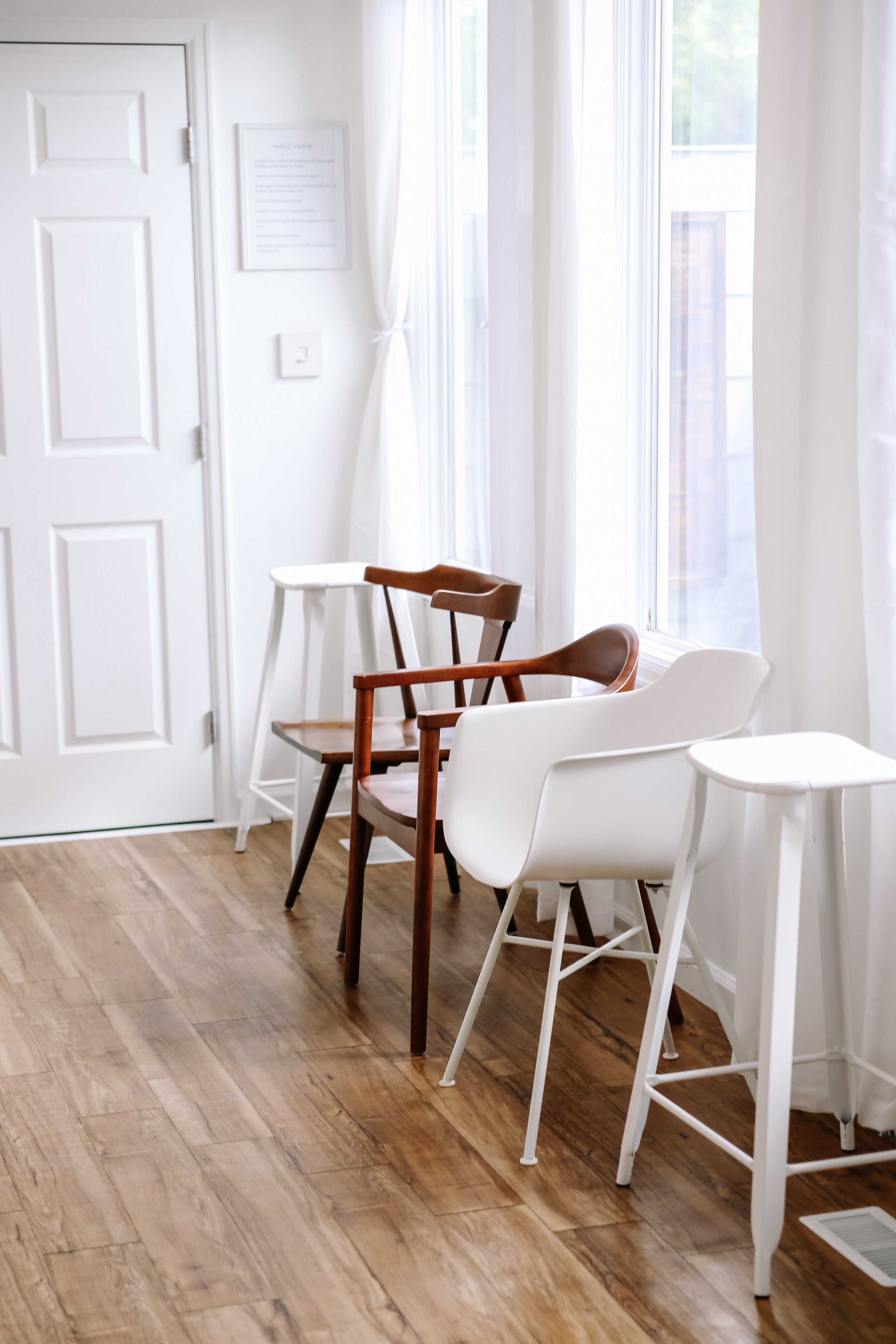 A row of chairs of different styles and colors, including white, wood, and brown, placed along a wall near a large window with white curtains in a room with wooden flooring.