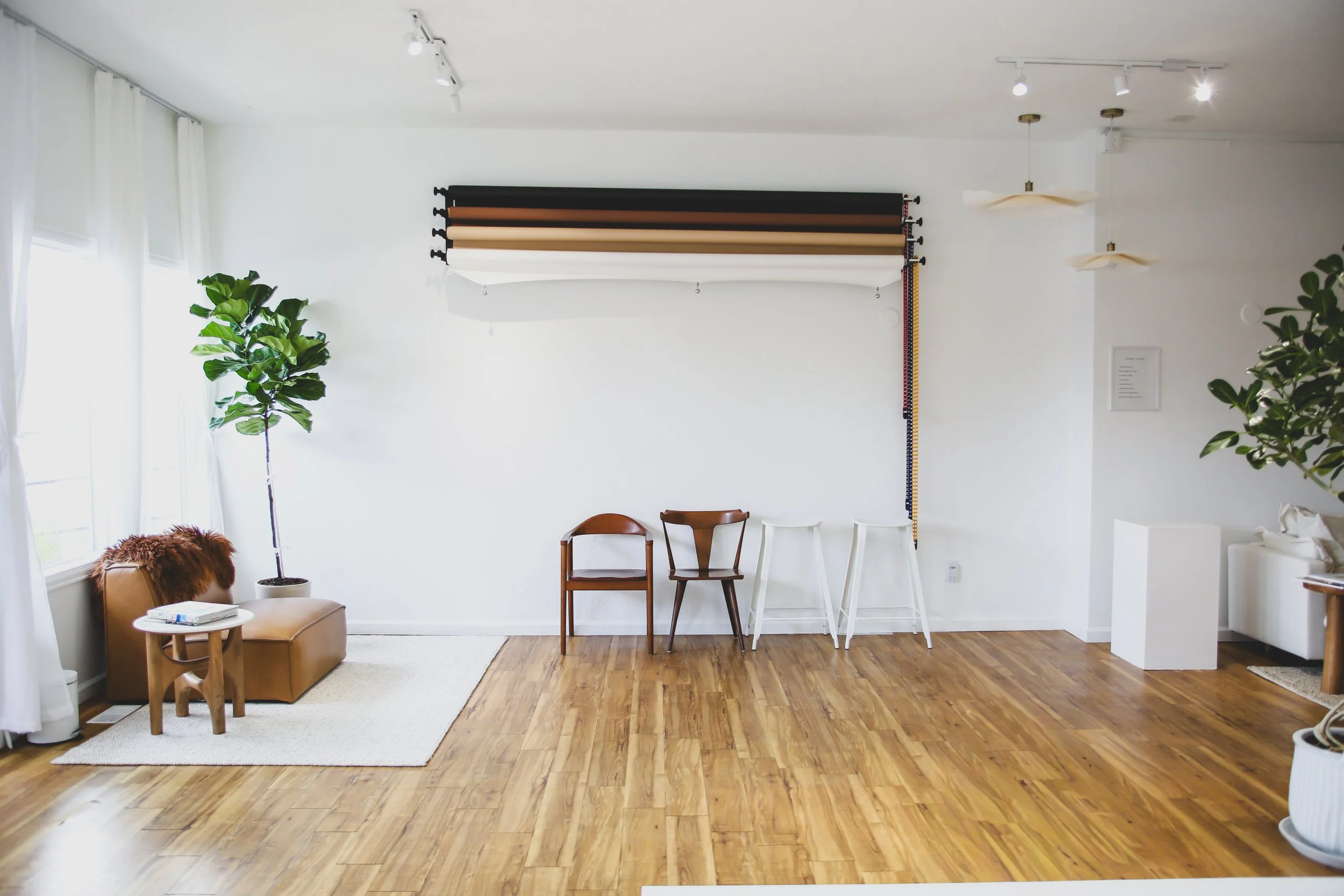 A bright, minimalist living space with white walls and hardwood floors. There is a large window to the left with white curtains, a small beige armchair with a furry pillow, and a white side table. Two green plants are present, one near the window and another on the right side of the room. Four wooden chairs are placed against the wall, and a set of rolls of wallpaper or fabric is mounted on the wall above the chairs. The ceiling has adjustable spotlights and pendant light fixtures.