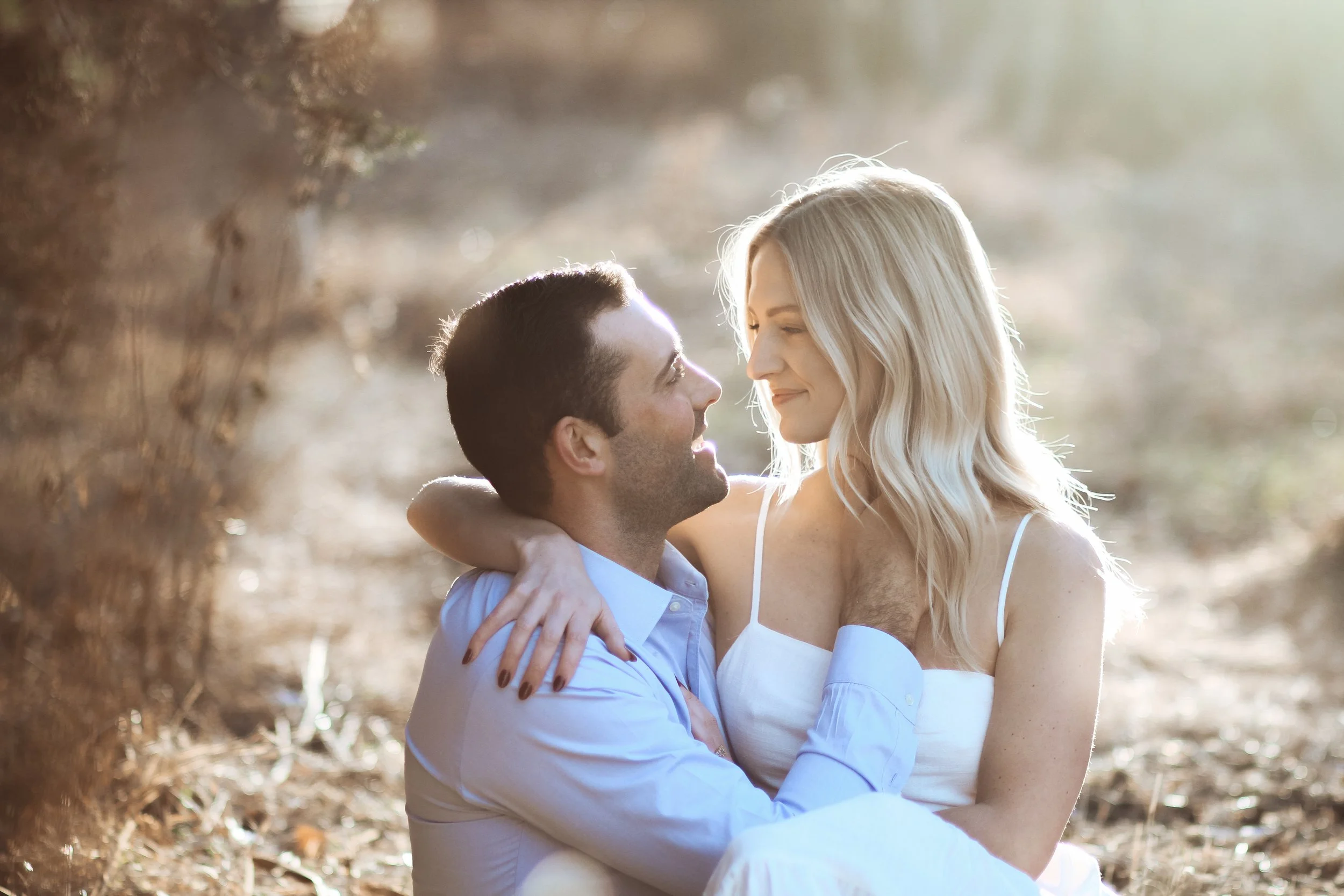 A couple sitting closely outdoors, smiling and looking at each other with sunlight behind them.