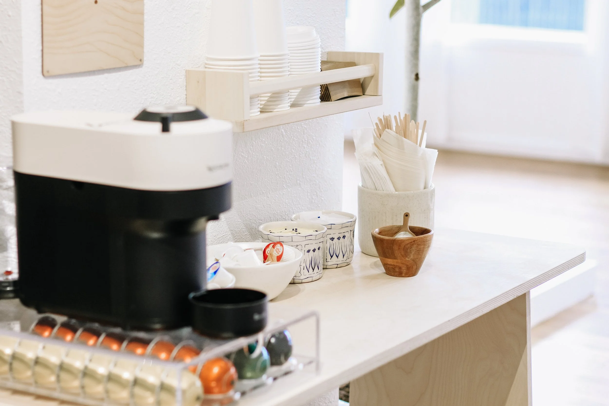 Coffee station with cups, bowls, and utensils on a white countertop, with a coffee maker in the foreground and natural light coming through a window.