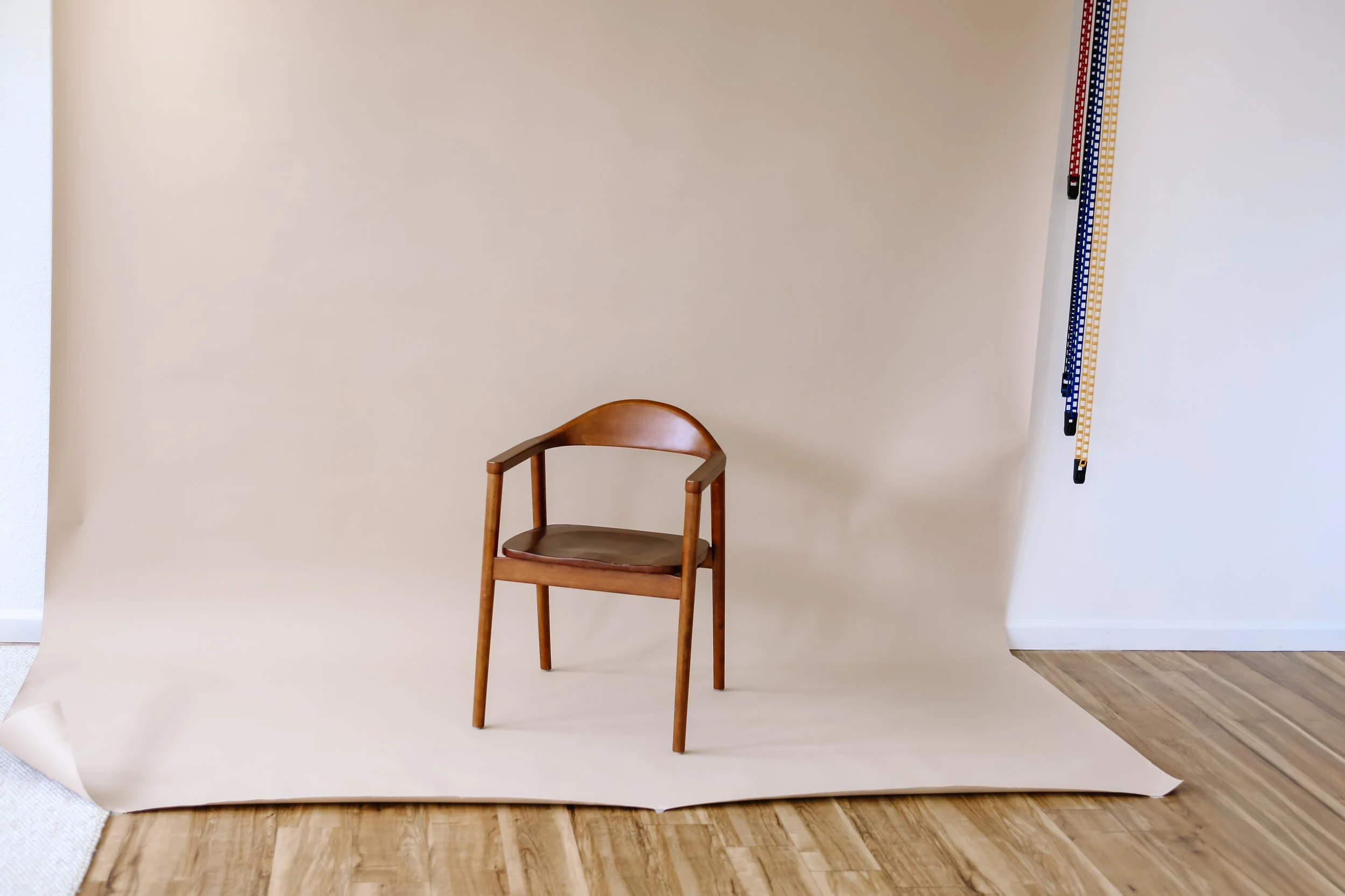 Photo of a simple wooden chair with a curved backrest positioned slightly in front of a plain beige backdrop supported by a paper roll stand.
