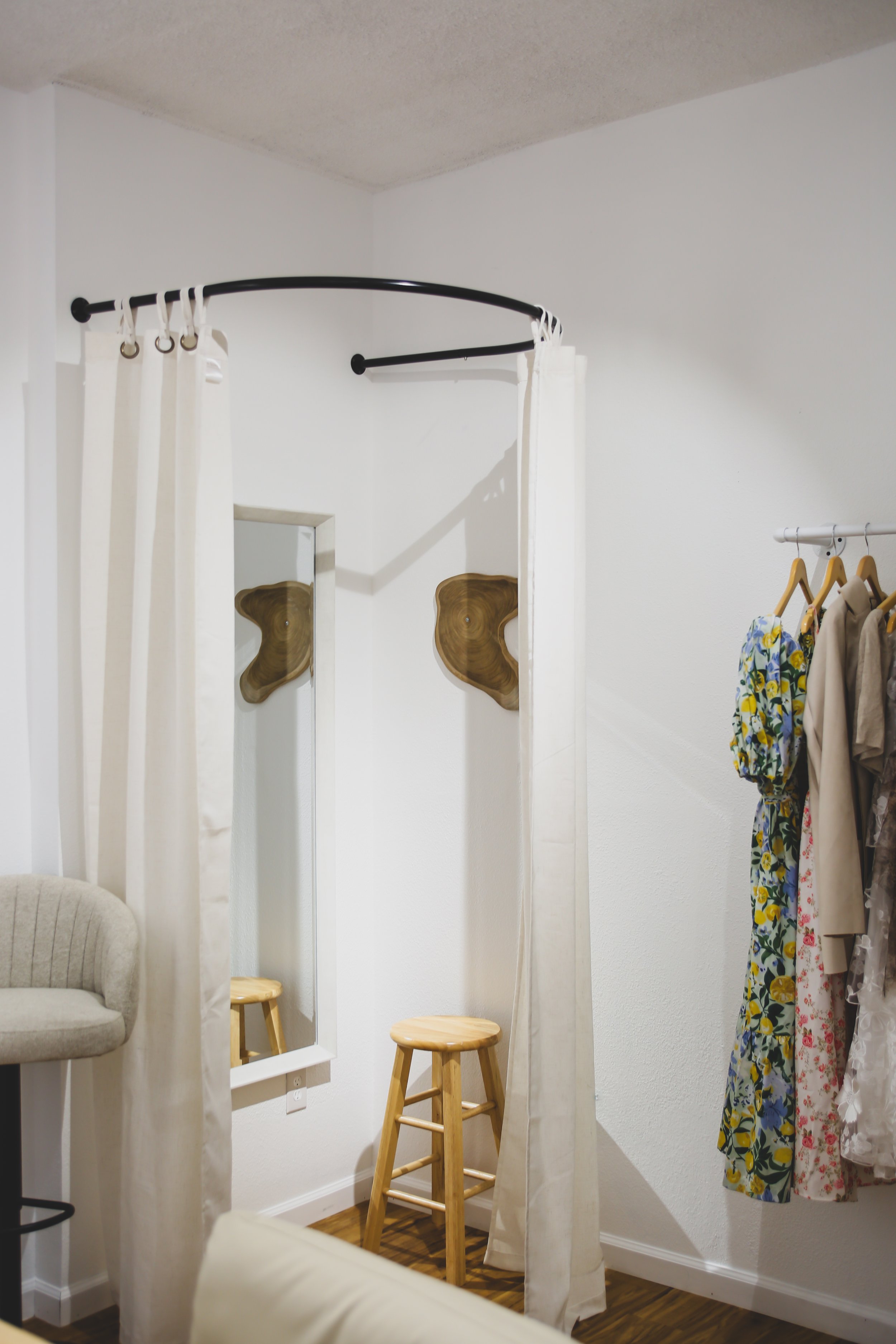 A fitting room area with a full-length mirror, wooden stool, white curtains, and clothing hanging on a rack.