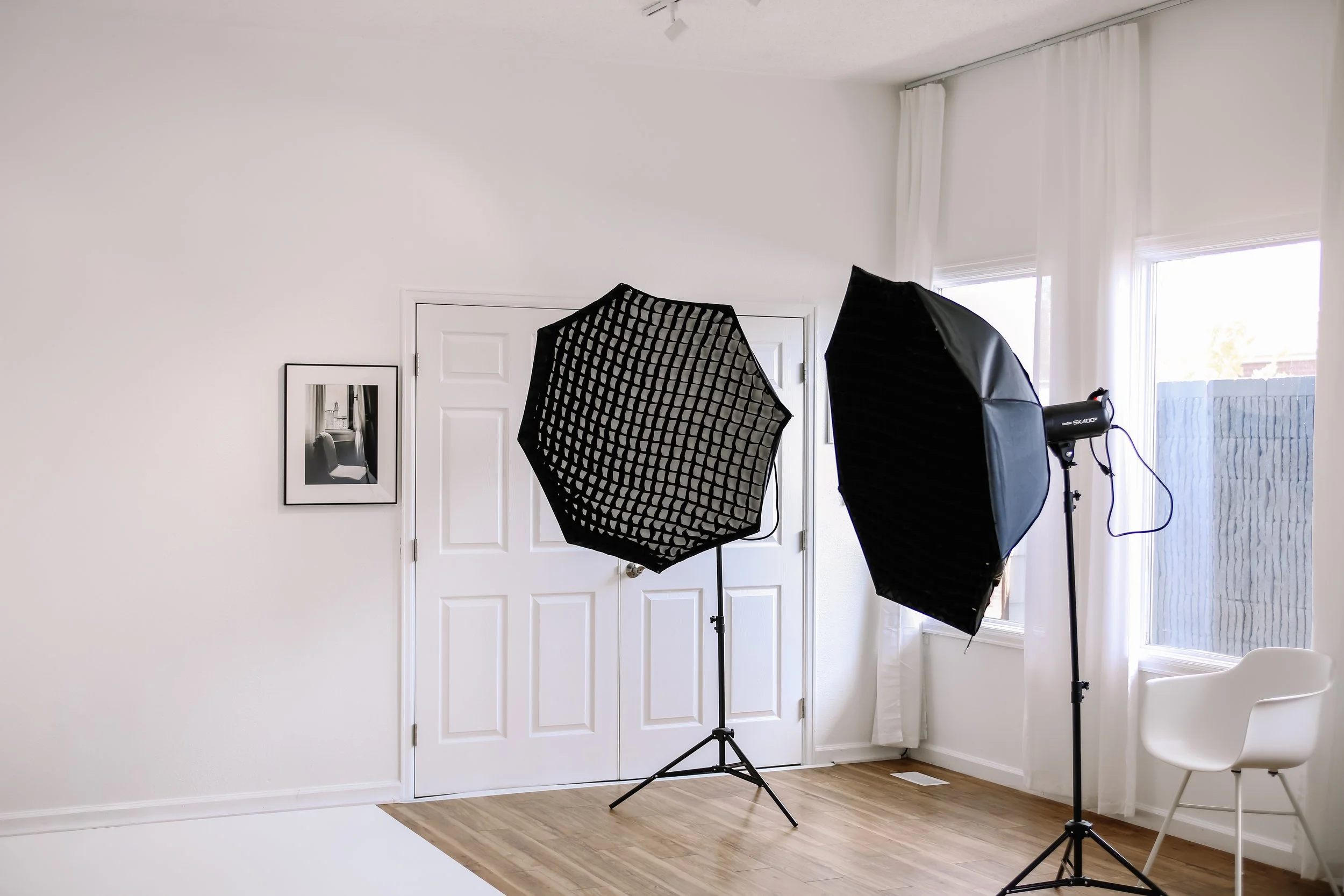 Photography studio setup with softbox lights, a white wall, framed black-and-white photograph, window with white curtains, white chairs, and hardwood floor.