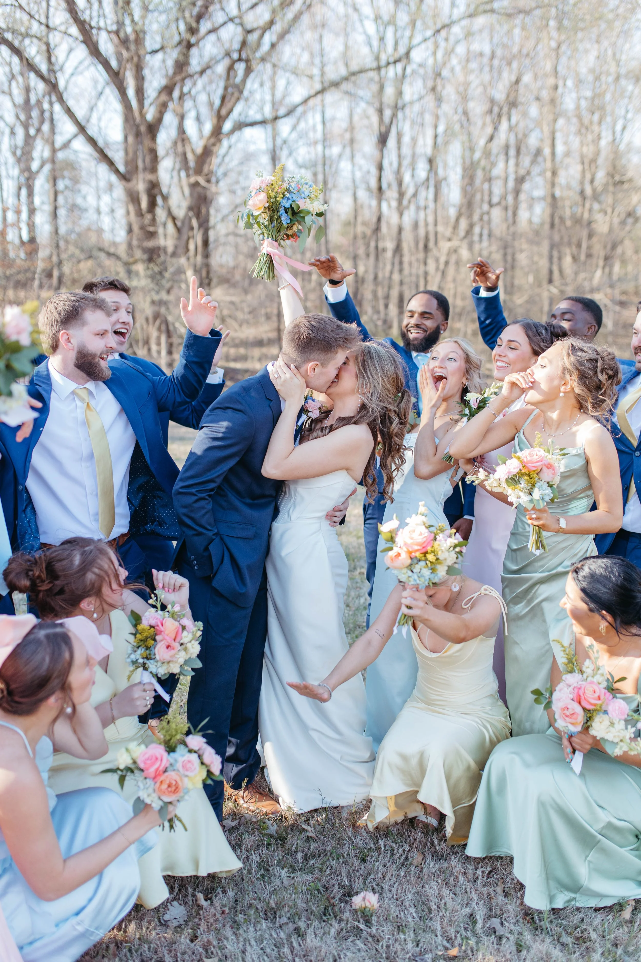A group of people celebrating a wedding outdoors, with a bride and groom kissing in the center, surrounded by friends and bridesmaids holding bouquets of flowers, most wearing pastel colors.