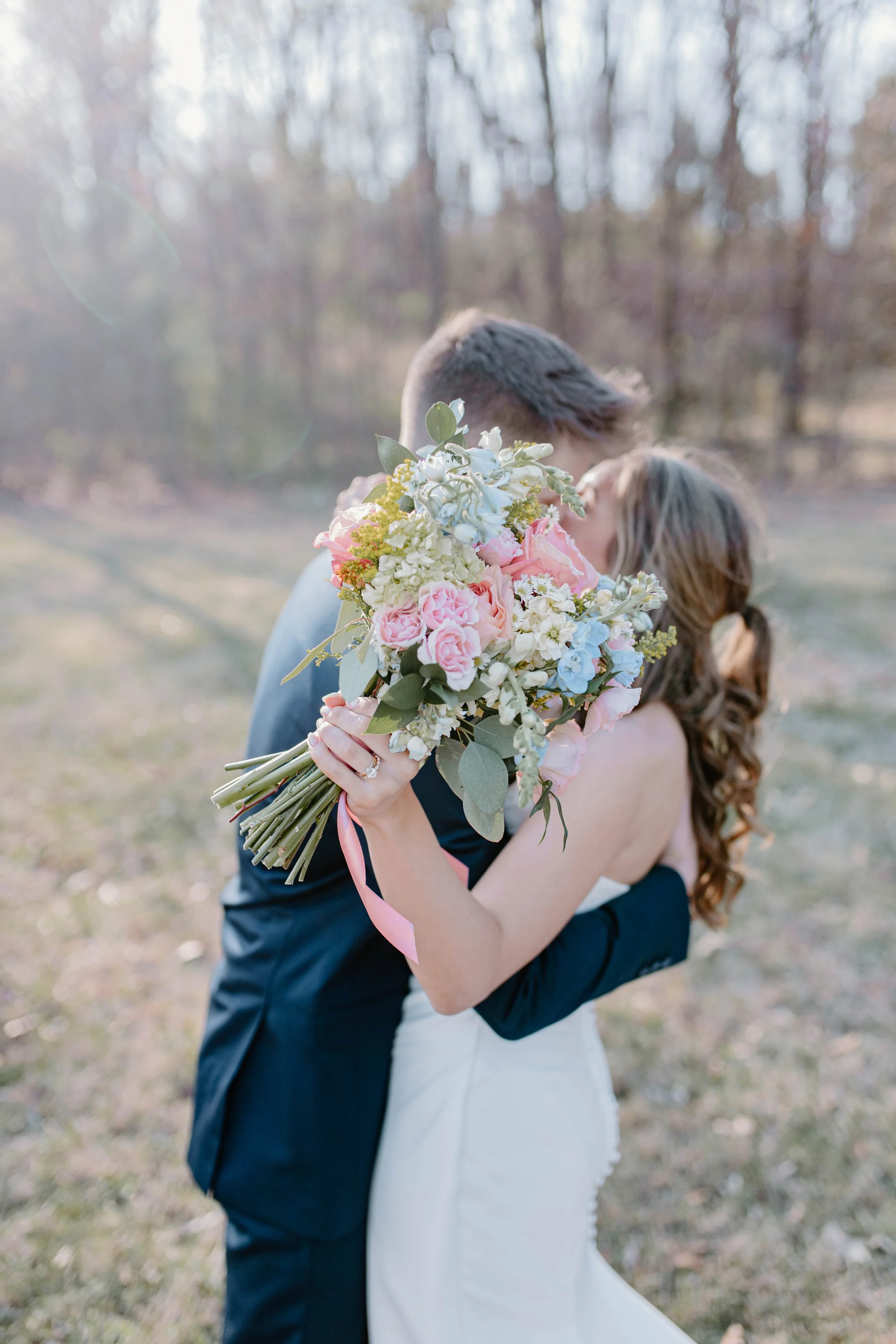A couple embracing outdoors, with the woman holding a bouquet of pink, white, and blue flowers.