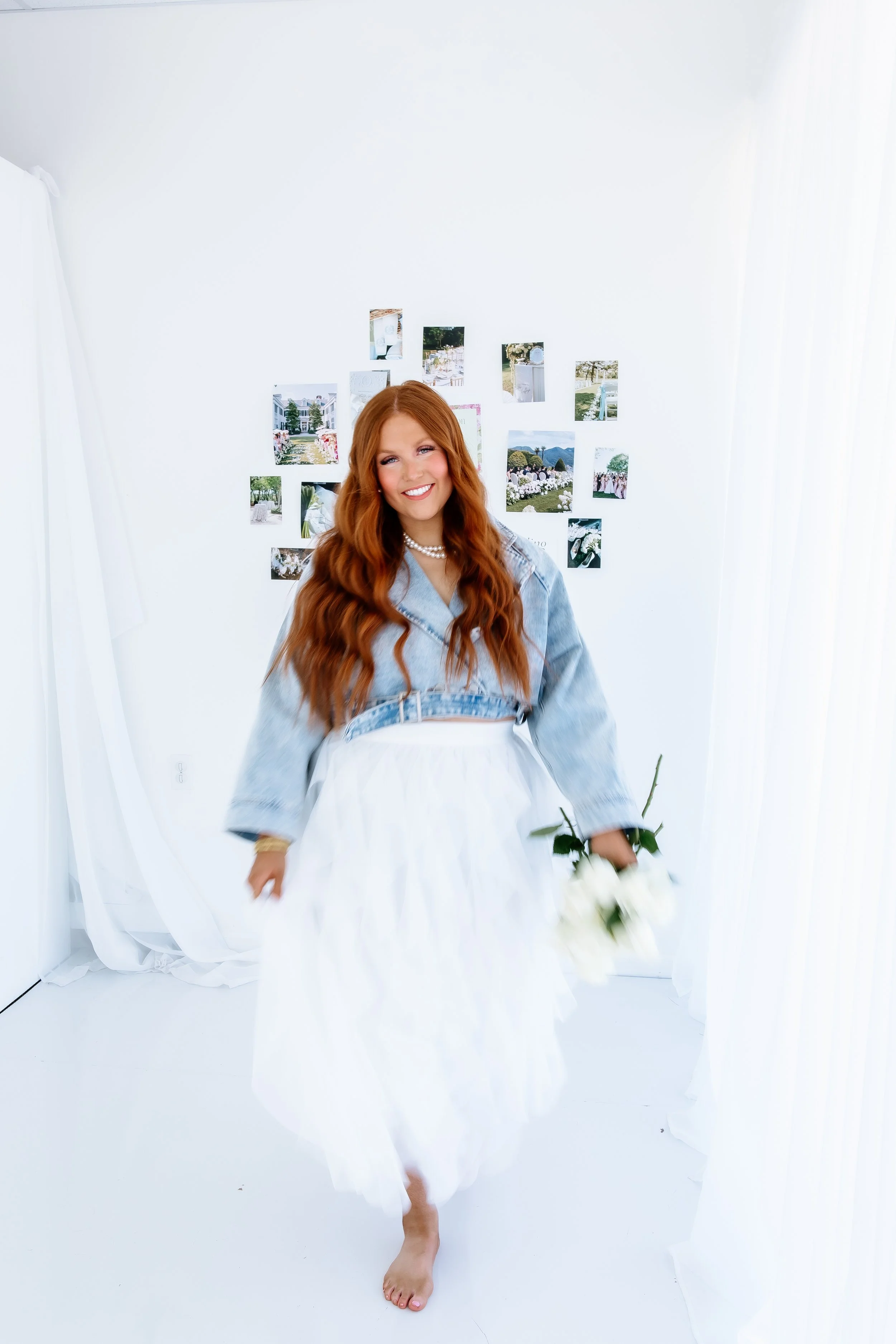A woman with long red hair smiling, holding white flowers, standing in a bright white room, wearing a denim jacket and white skirt.