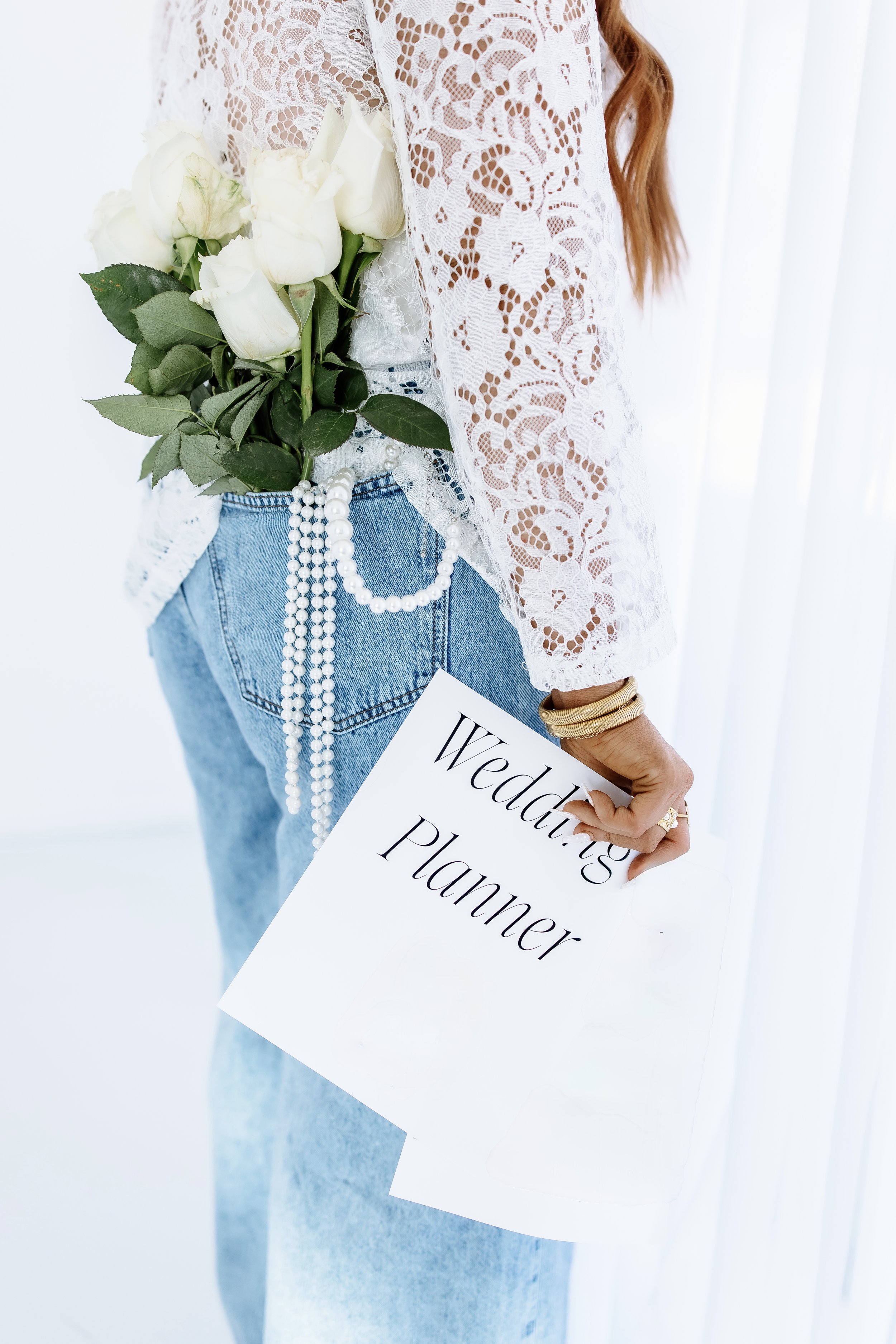 Woman holding a wedding planner booklet in her right hand, wearing a white lace top, light blue jeans, and gold bangles. She has a bouquet of white roses tucked into her back pocket, with a string of pearls hanging from her jeans.