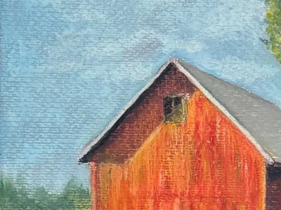 Close-up of a painted wooden barn with a gabled roof, set against a blue sky, with some green trees visible on the sides.