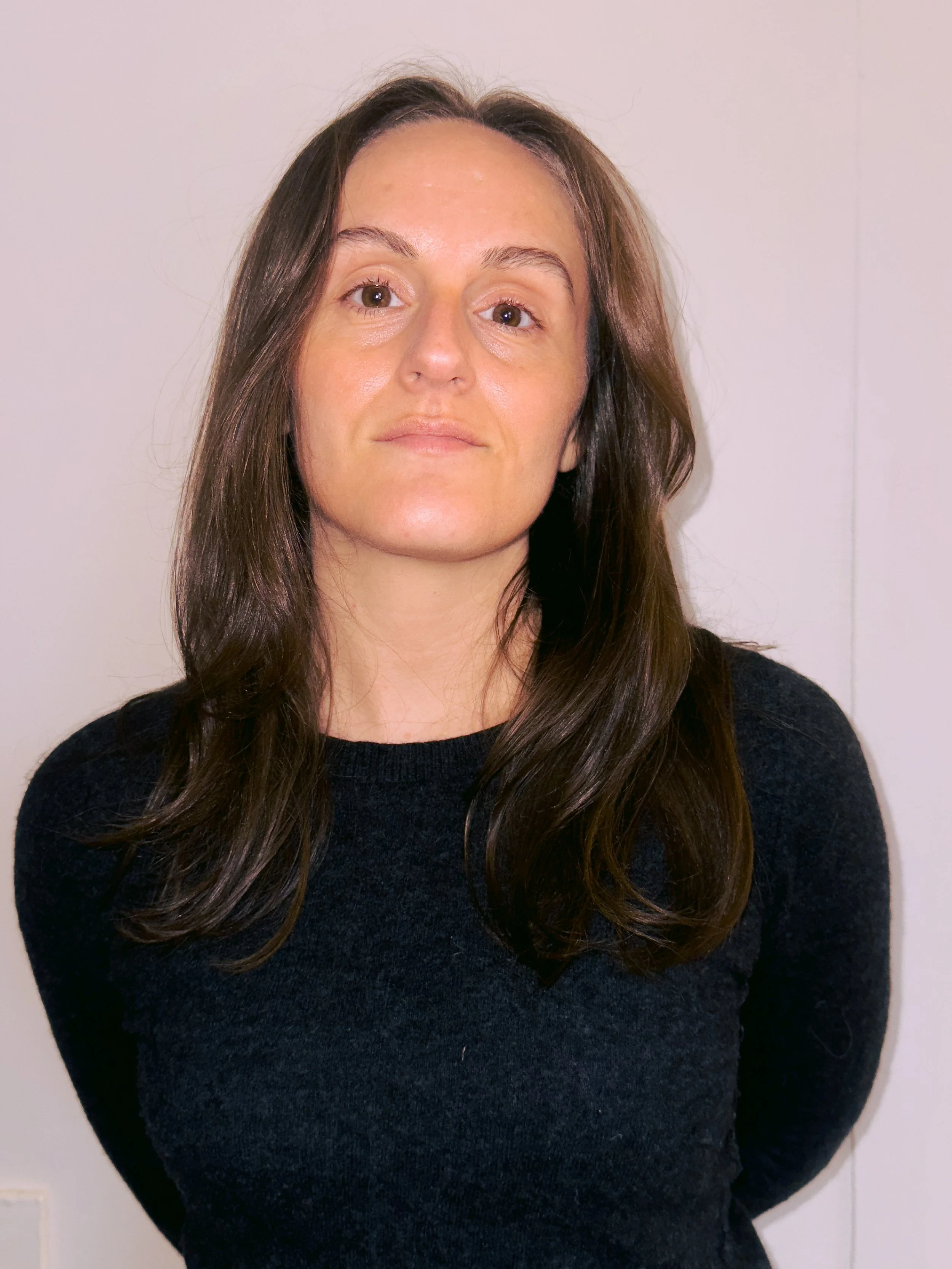 A woman with long, dark brown hair and light skin stands against a plain, off-white wall. She is looking directly at the camera with a neutral expression and is wearing a black sweater.