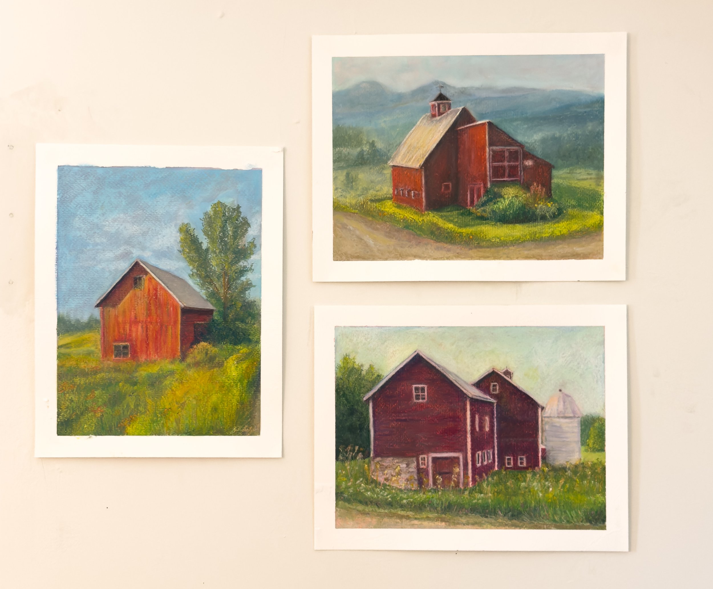 Three paintings of rural farmhouses on a white wall, depicting red barns surrounded by greenery and countryside.