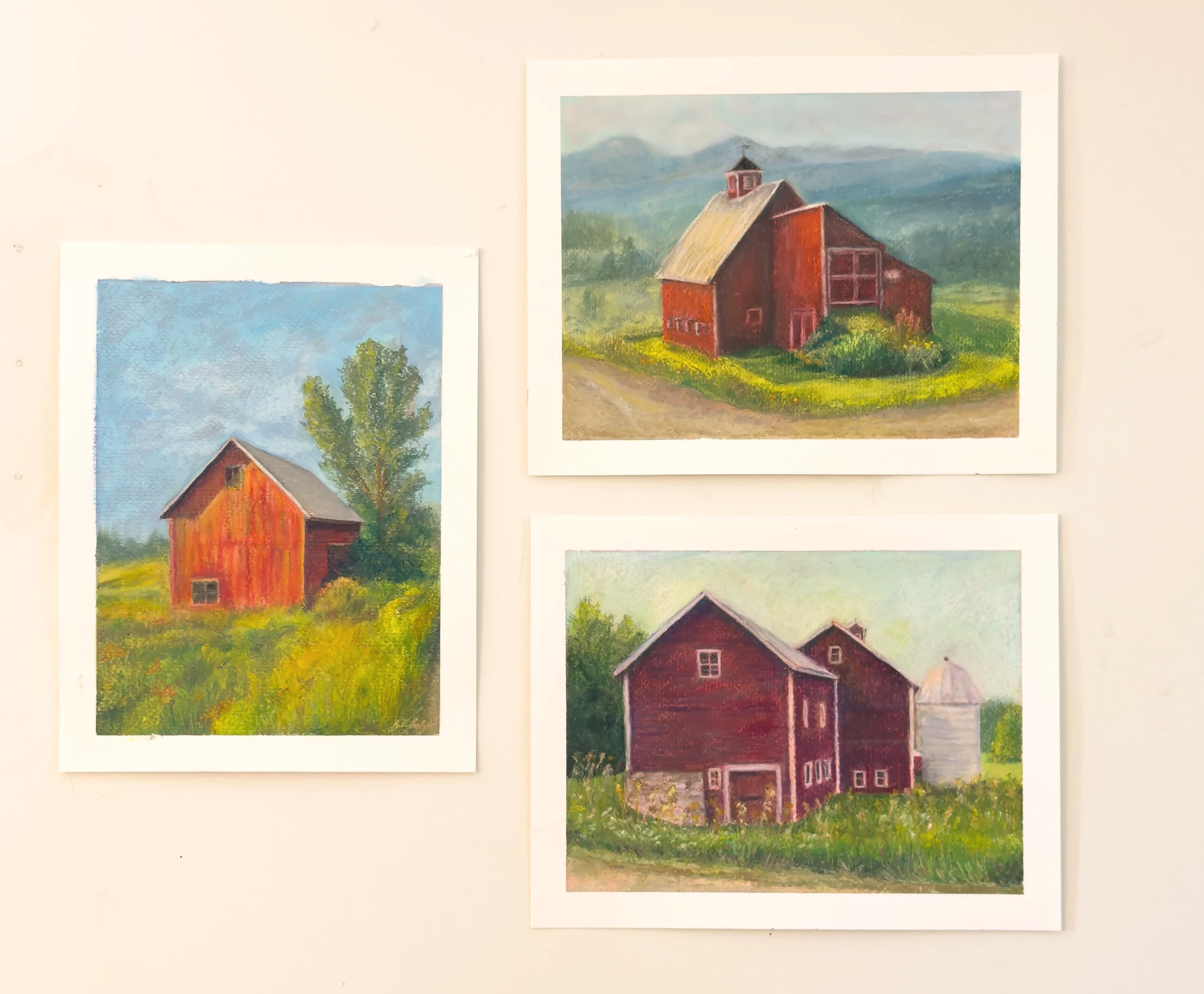 Three paintings of barns on a wall, each depicting red barns with grass and trees in a countryside setting.