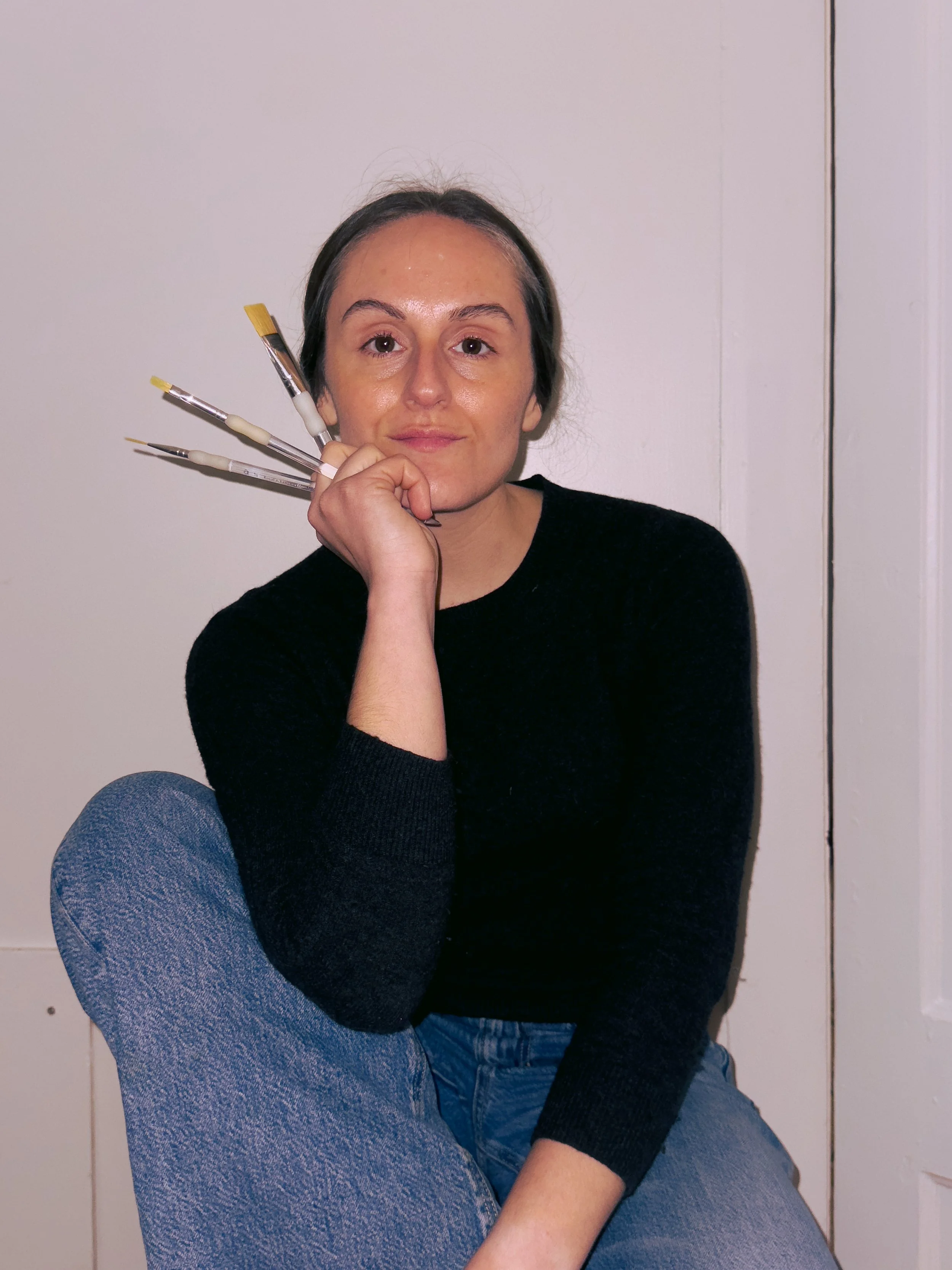 A woman sitting with her chin resting on her hand, holding four paintbrushes near her face, wearing a black sweater and blue jeans, sitting against a white wall.