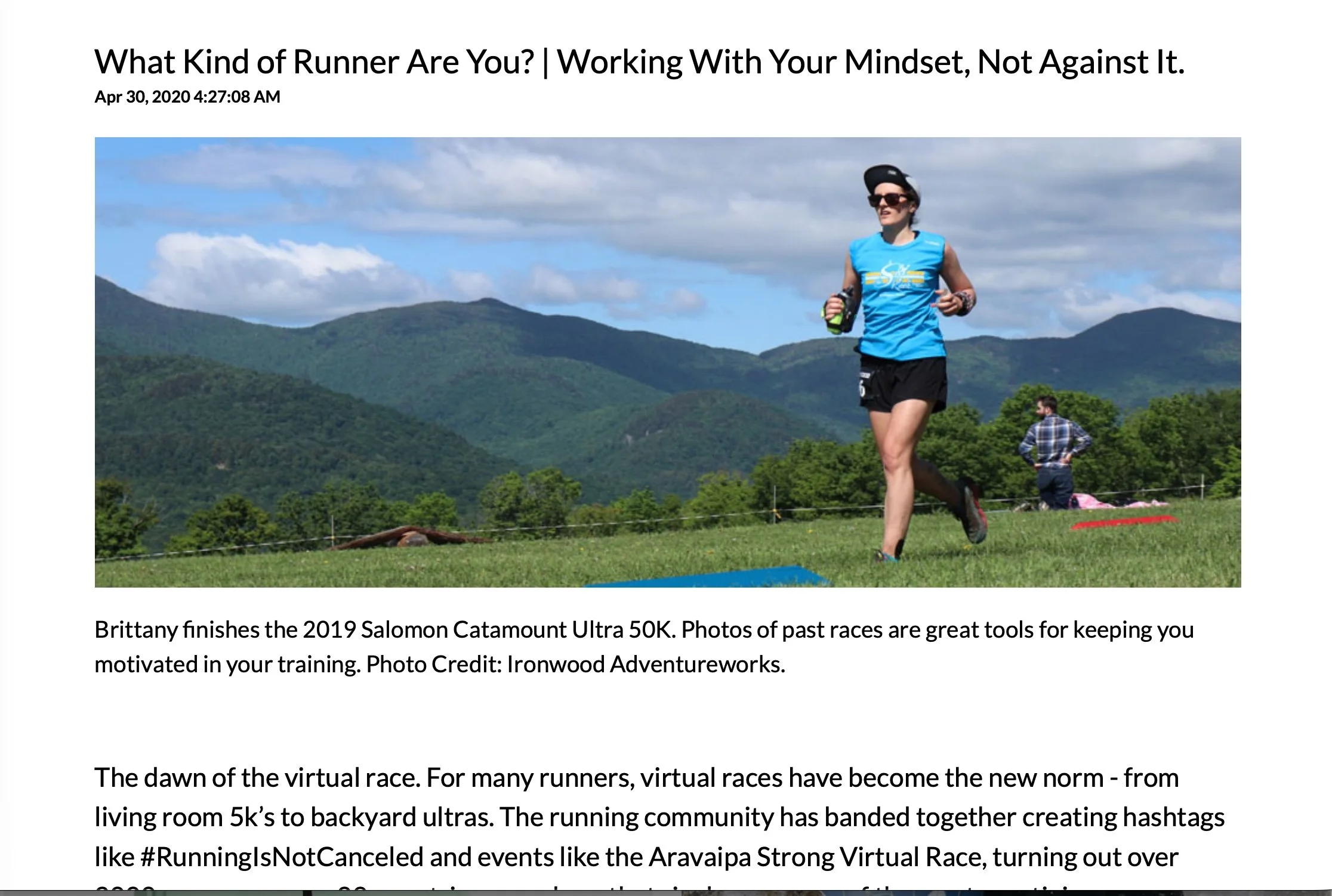 A woman wearing a blue shirt, black shorts, sunglasses, and a hat running outdoors on a grassy field with mountains and blue sky in the background.