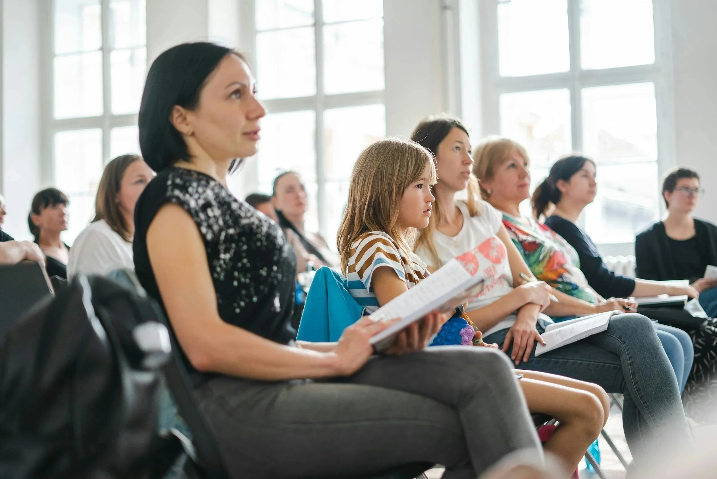 Group of women and a girl attending a seminar or conference in a bright room with large windows, sitting on chairs with notebooks or papers.