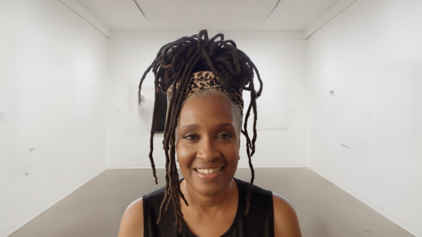 Woman with dreadlocks styled up, wearing a leopard print headband, smiling in an art gallery with white walls.