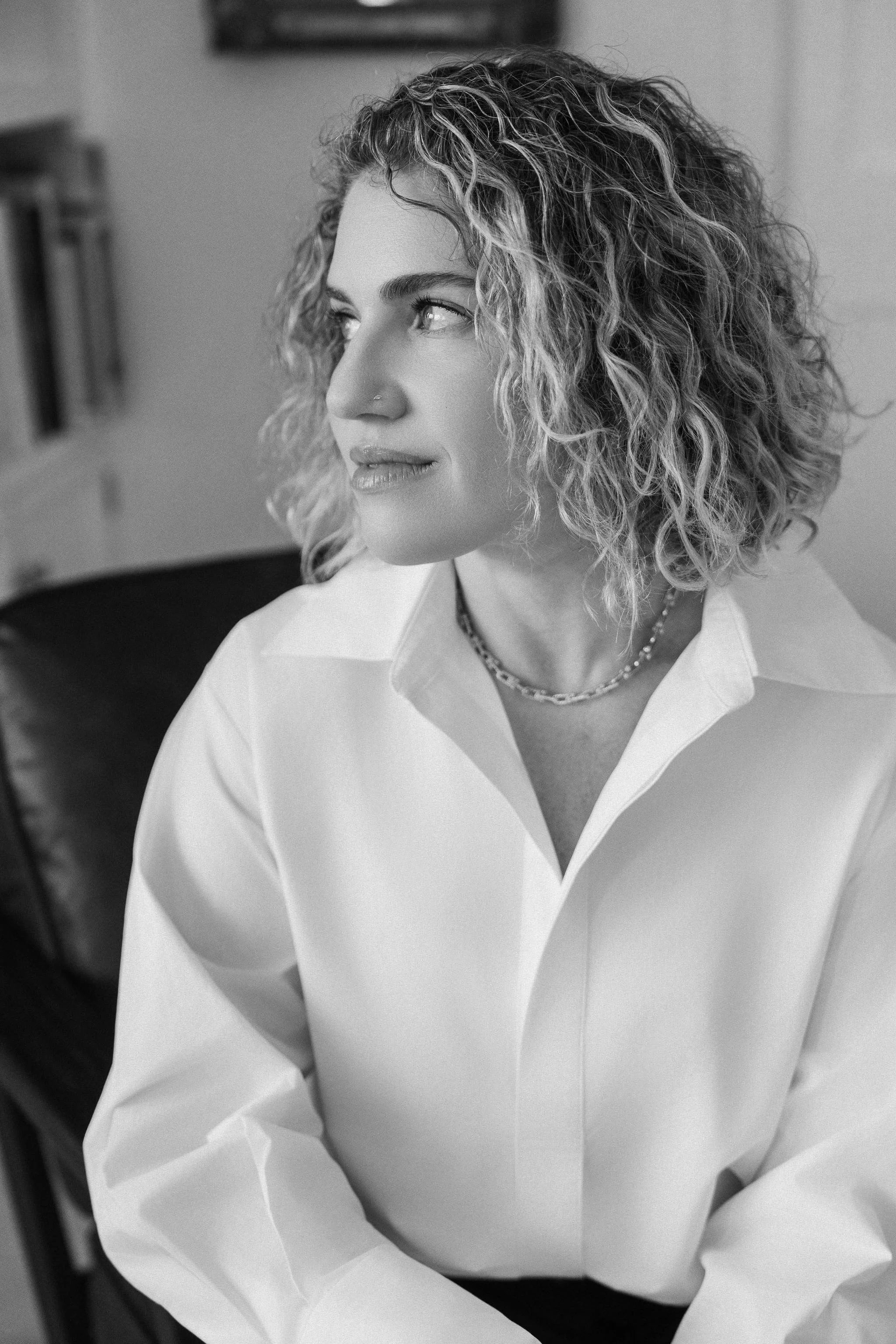 Black and white photo of a woman with curly hair, wearing a white shirt and a necklace, looking to the side.