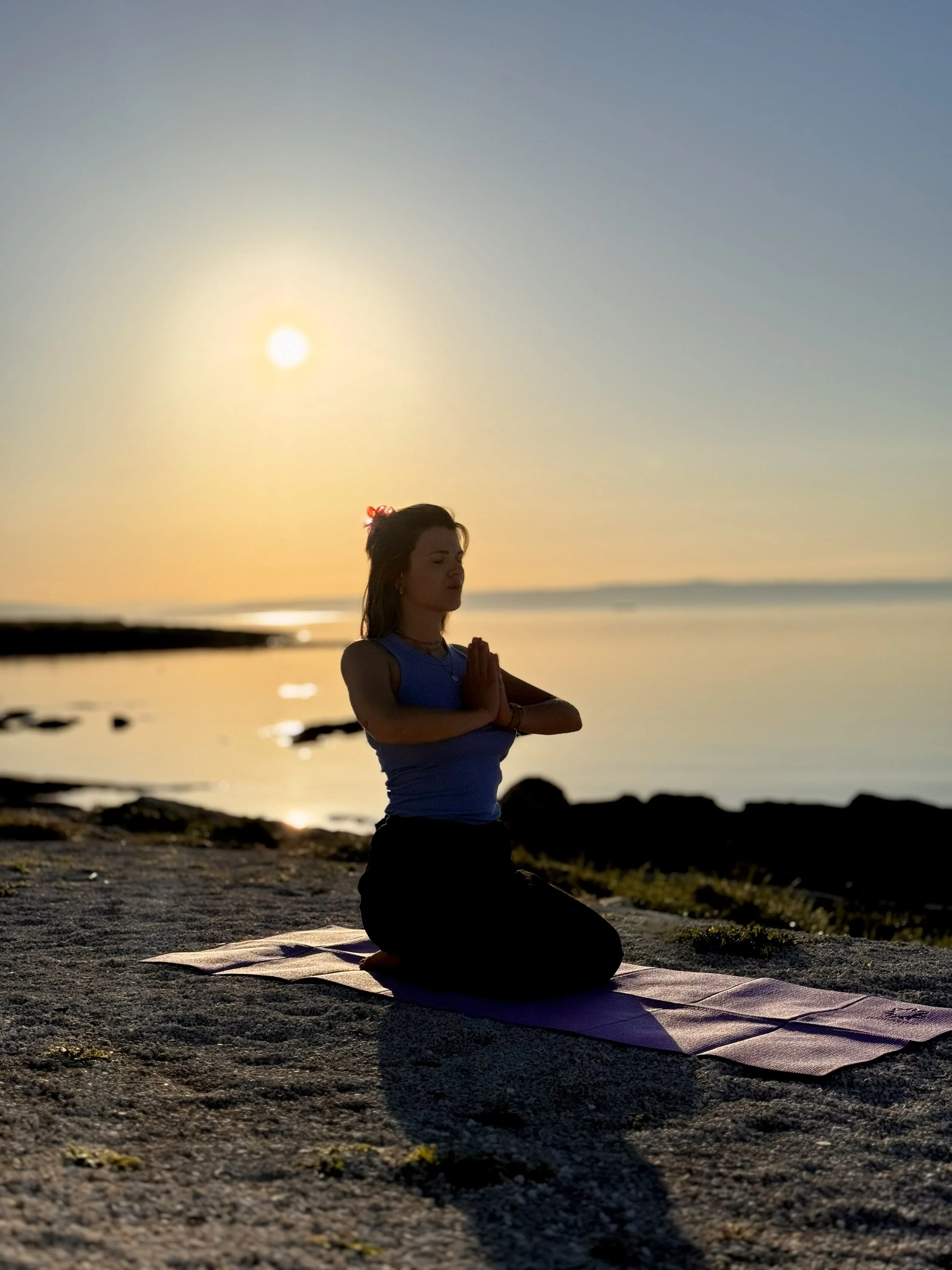 Frau, die im Lotussitz auf einer Yogamatte am Strand bei Sonnenuntergang sitzt und meditiert.