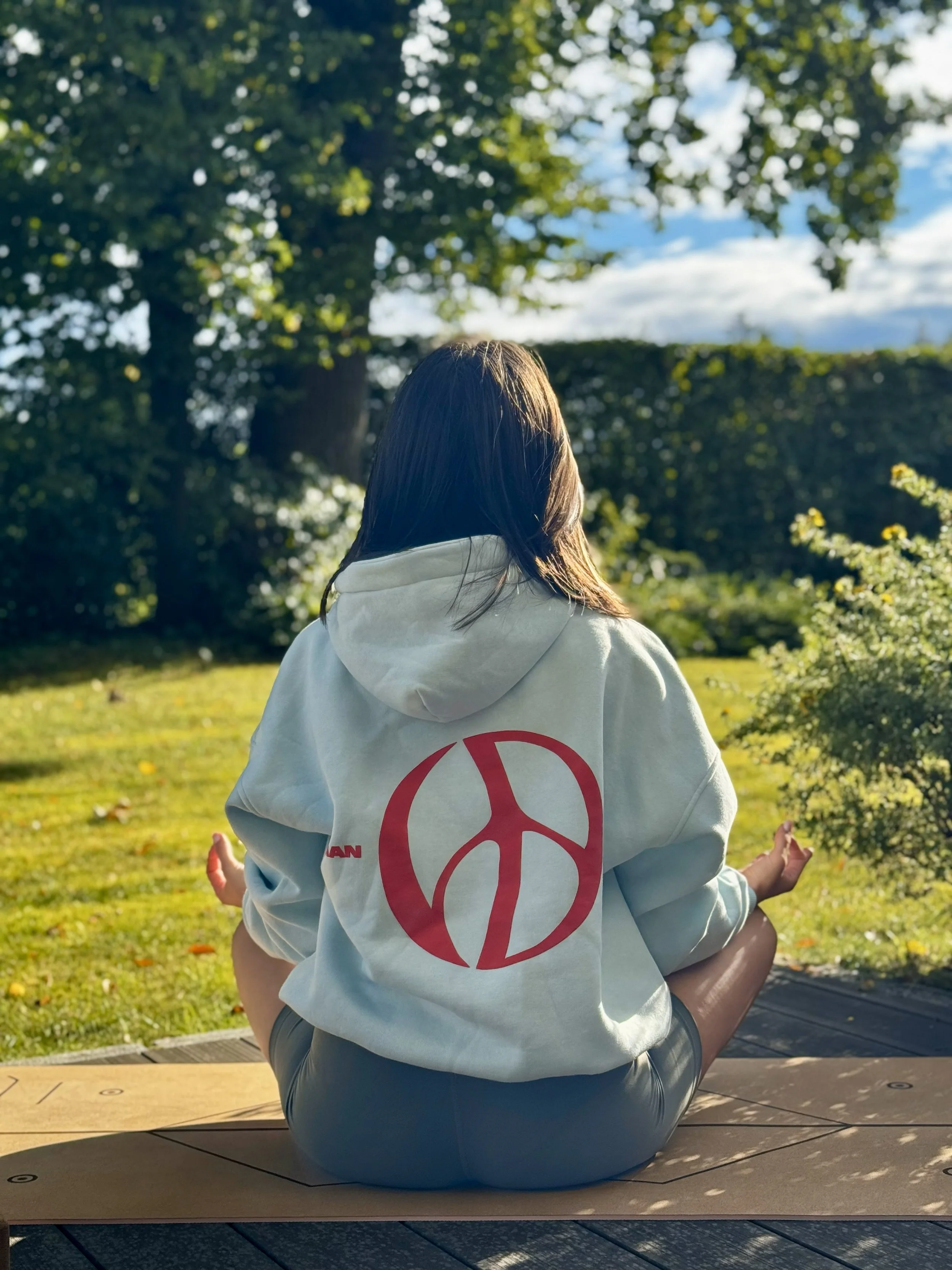 Eine Person sitzt im Schneidersitz auf einer Holzplattform in einem Garten, trägt ein helles Sweatshirt mit einem roten Peace-Zeichen auf dem Rücken, blickt auf eine grüne Hecke und Bäume bei Sonnenschein.