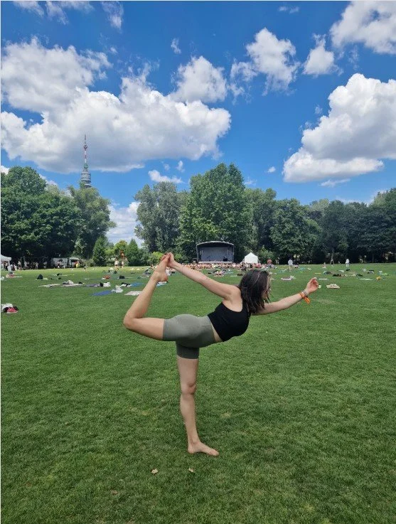 Eine Frau macht Yoga im Freien auf einer grünen Wiese, umgeben von Bäumen, mit einem blauen Himmel und weißen Wolken.