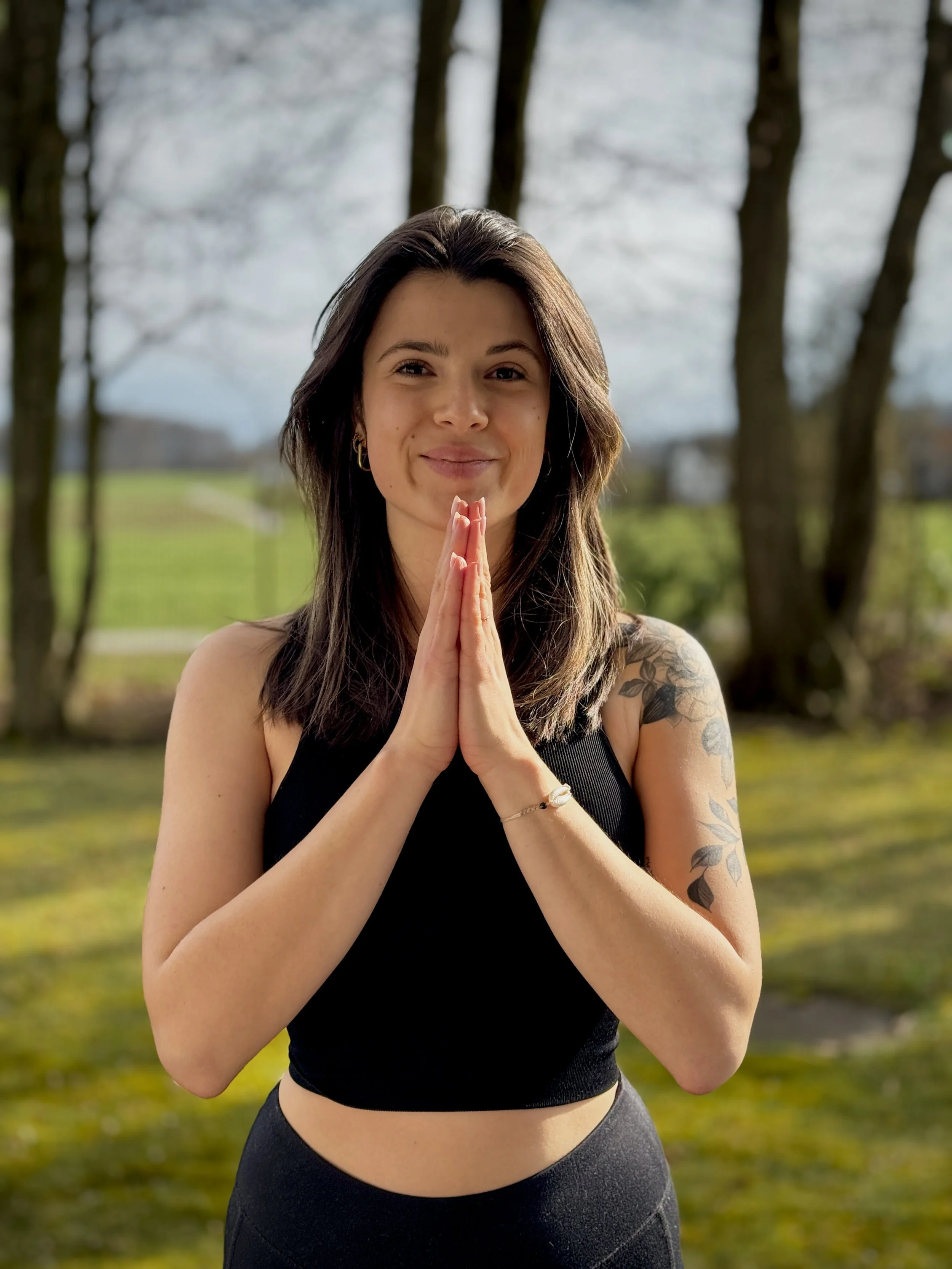 Eine junge Frau mit dunklen Haaren, steht im Freien vor Bäumen. Sie hält die Hände in Gebetsposition vor der Brust und lächelt.