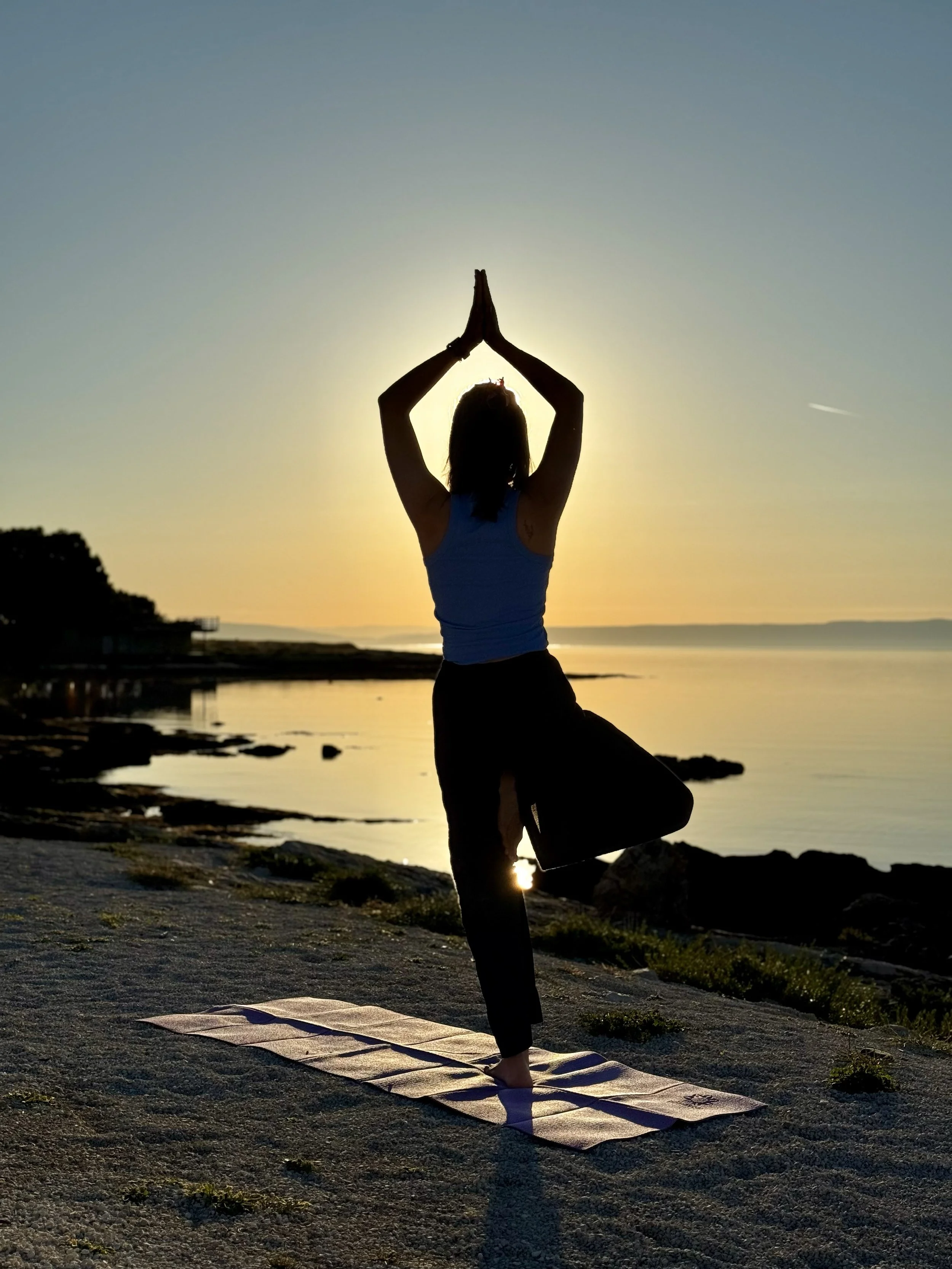 Eine Frau macht Yoga am Strand bei Sonnenuntergang, steht in der Baumposition mit den Händen über dem Kopf, auf einer Matte.