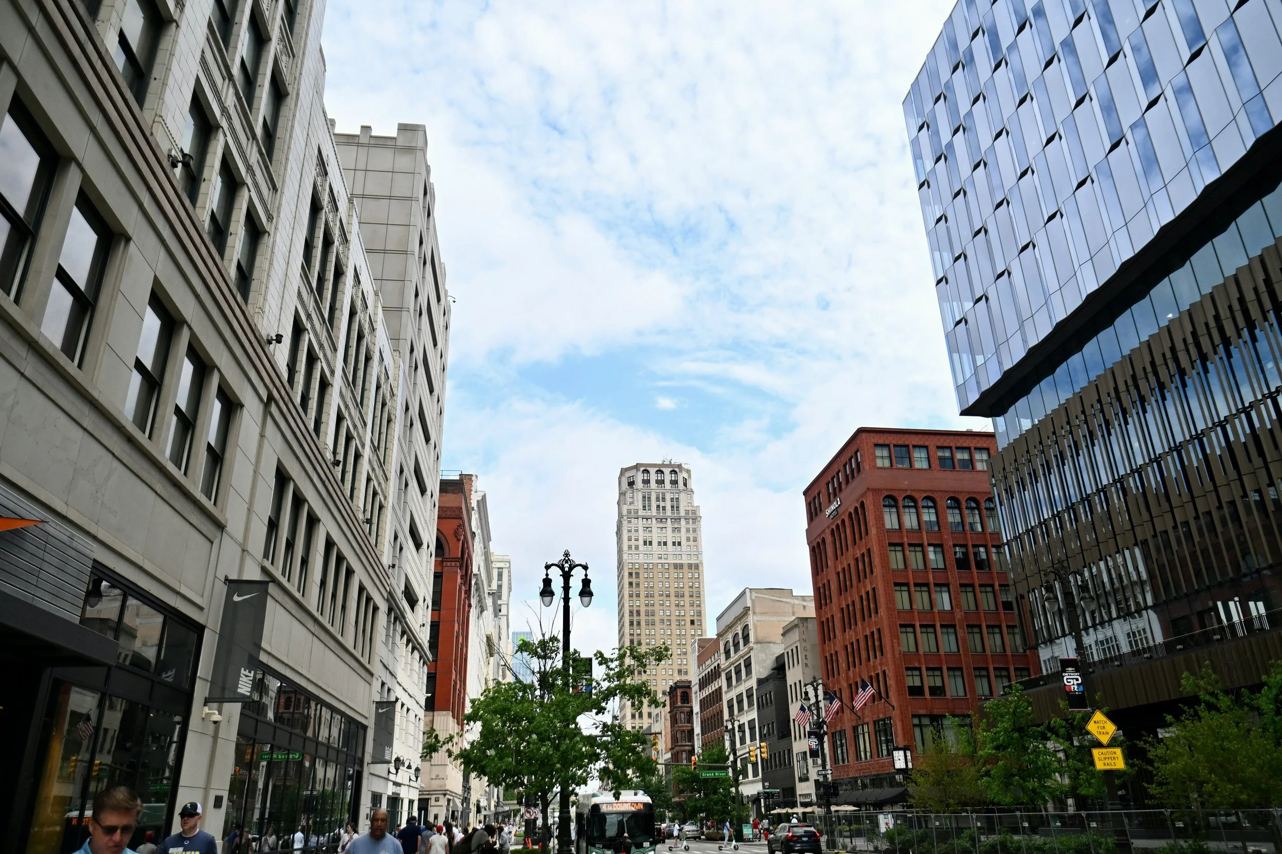 A city street scene with tall buildings on both sides, pedestrians on the sidewalk, and a city bus. The sky is partly cloudy.