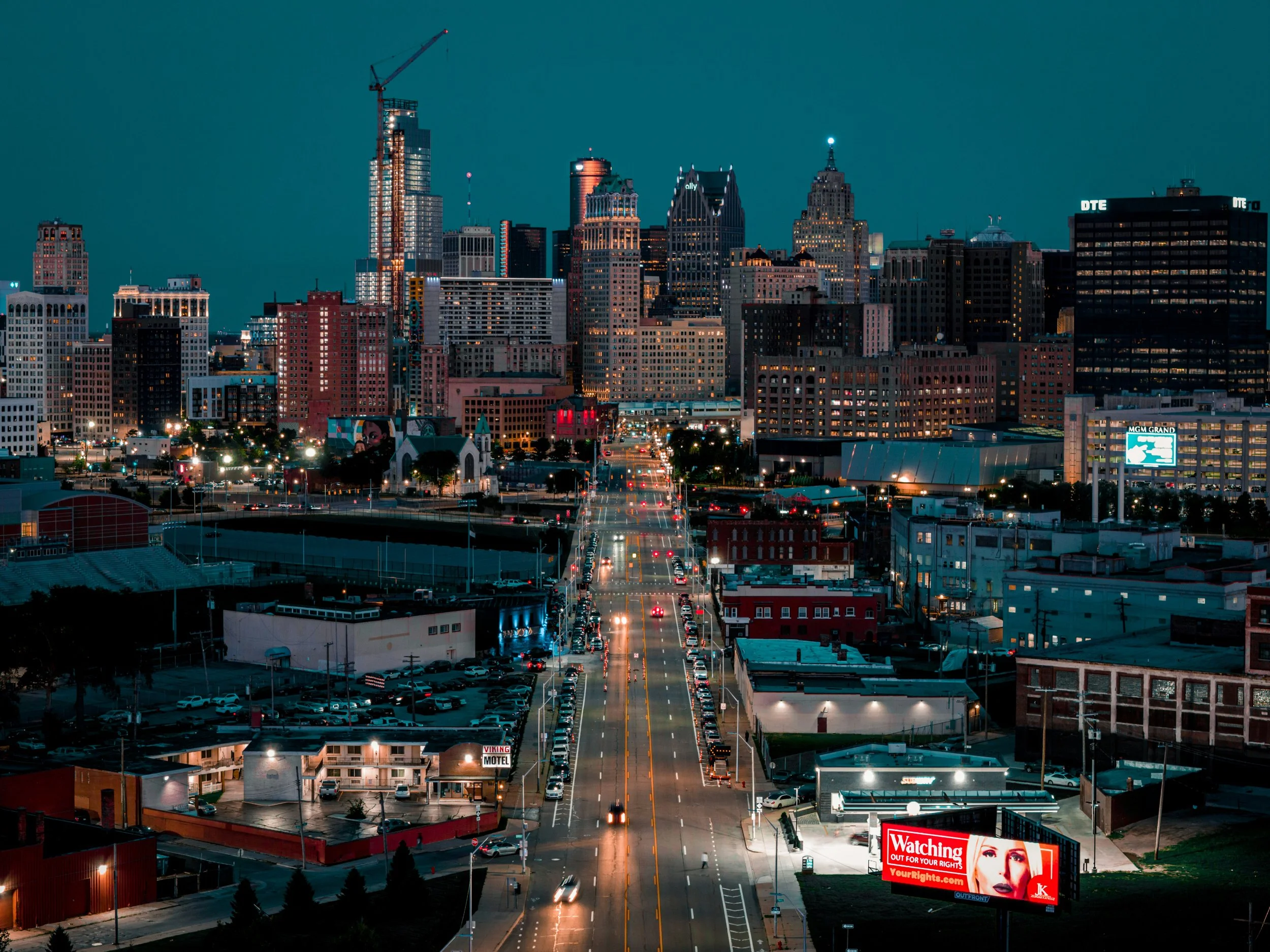 Night view of a city skyline with tall skyscrapers, some under construction, illuminated window lights, and a main street with moving vehicles, billboards including one that says 'Watching Out For Your Rights' and 'yourrights.com'.
