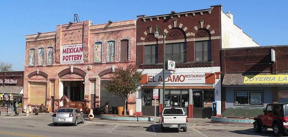 Street view of a small city block with various storefronts, including a pottery shop, a restaurant, and a jewelry store. Cars are parked in front, and there is a small tree and a decorative fountain in the scene.