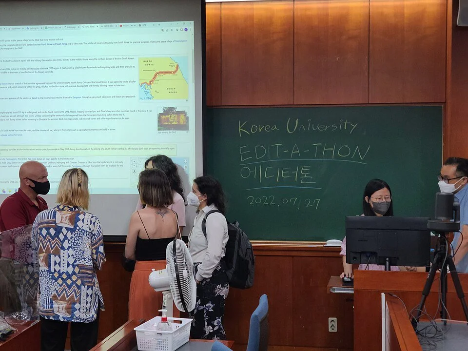 Group of students and a teacher in a classroom participating in an editing event at Korea University on July 27, 2022, with a chalkboard and a large screen project showing a map and text in the background.