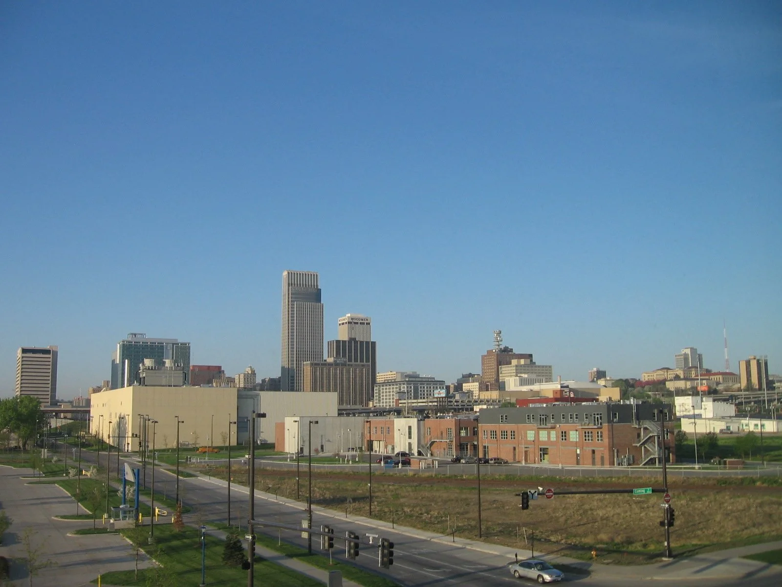 City skyline with modern high-rise buildings under a clear blue sky, foreground includes streets, sidewalk, and some smaller buildings.