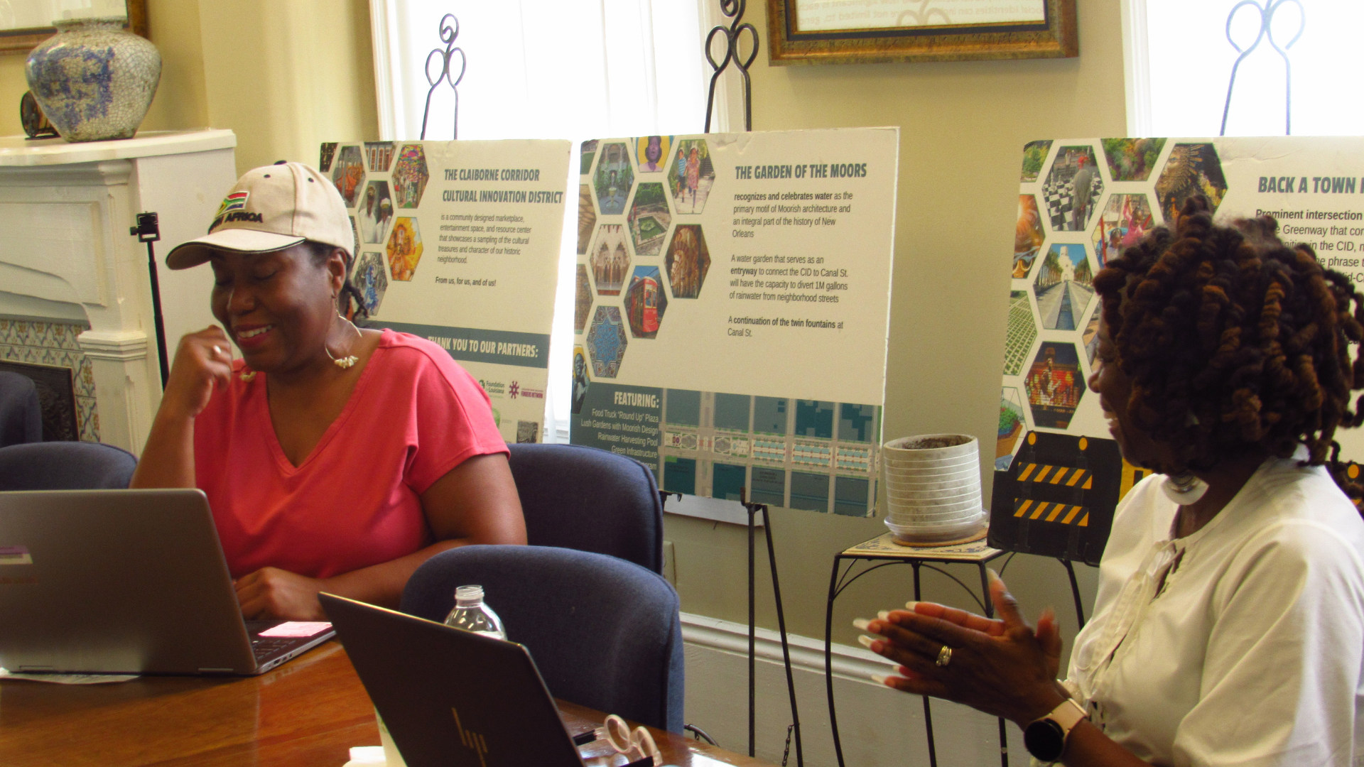 Two women sitting at a table with laptops, engaged in conversation. Behind them are display boards with colorful information and images, suggesting a presentation or community event.