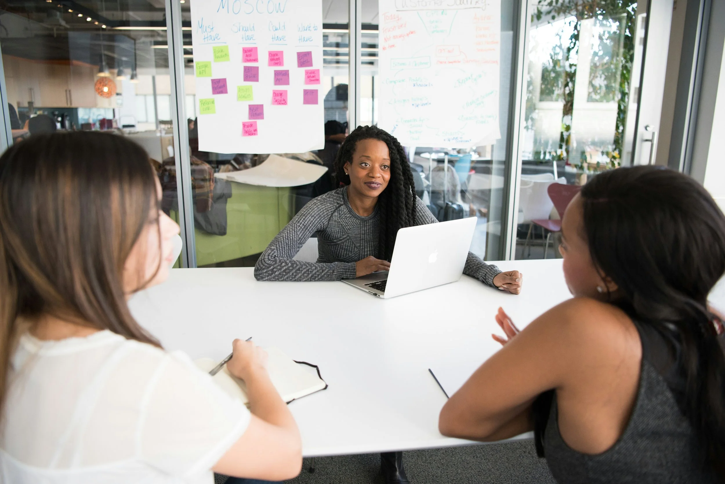 A woman with long, curly hair sitting at a table with three other women in a modern office setting. Behind her, glass walls display colorful post-it notes and handwritten notes on large sheets of paper. The women are engaged in a discussion, with one woman taking notes in a notebook.