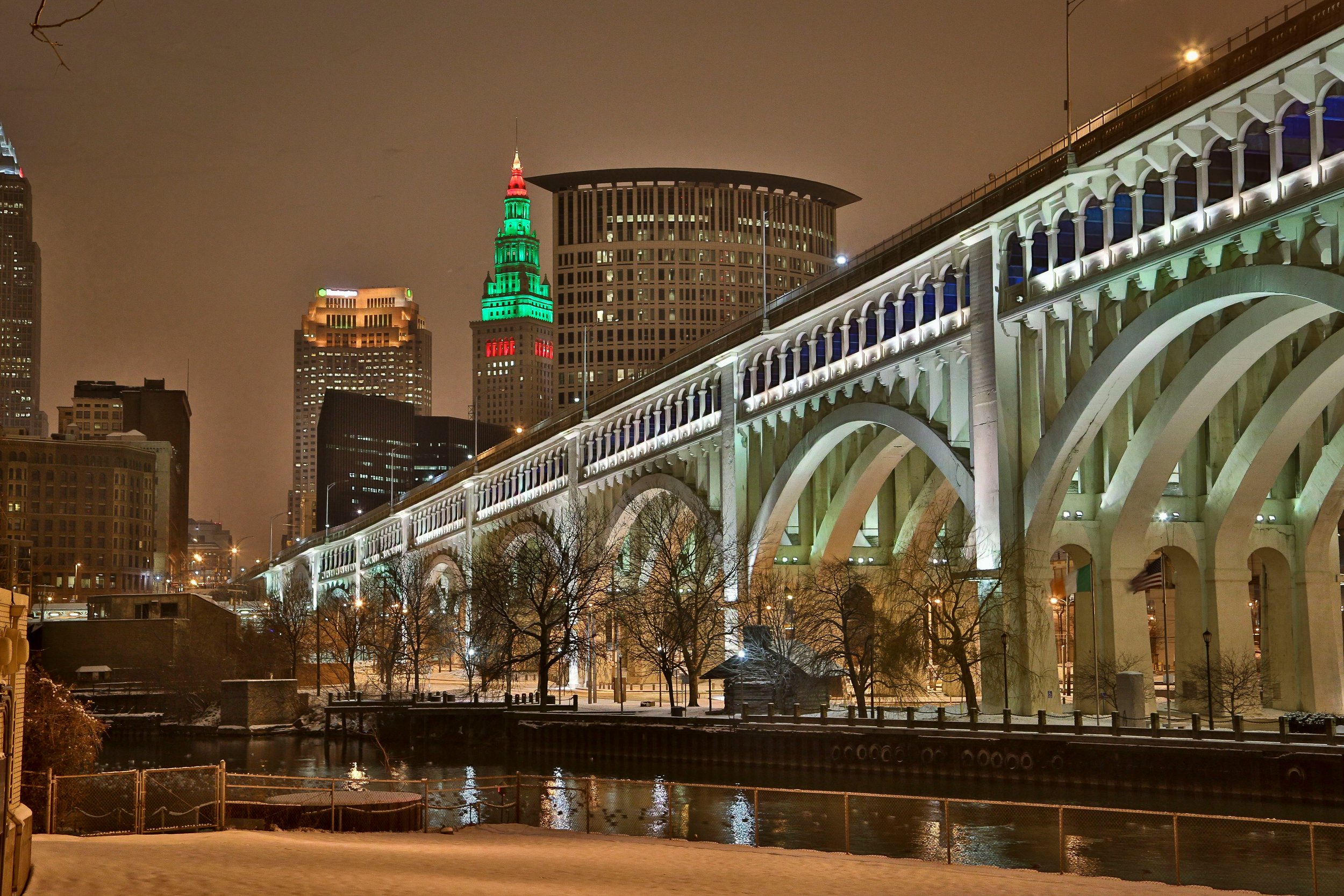 Nighttime cityscape with illuminated buildings, a bridge, and a river in Cleveland, Ohio. One building is lit with green and red lights in the shape of a Christmas tree.