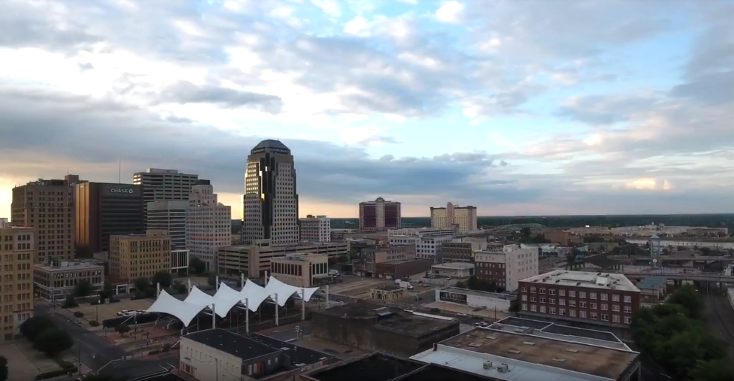 A city skyline at dusk with tall buildings, some tents in the foreground, and a partly cloudy sky