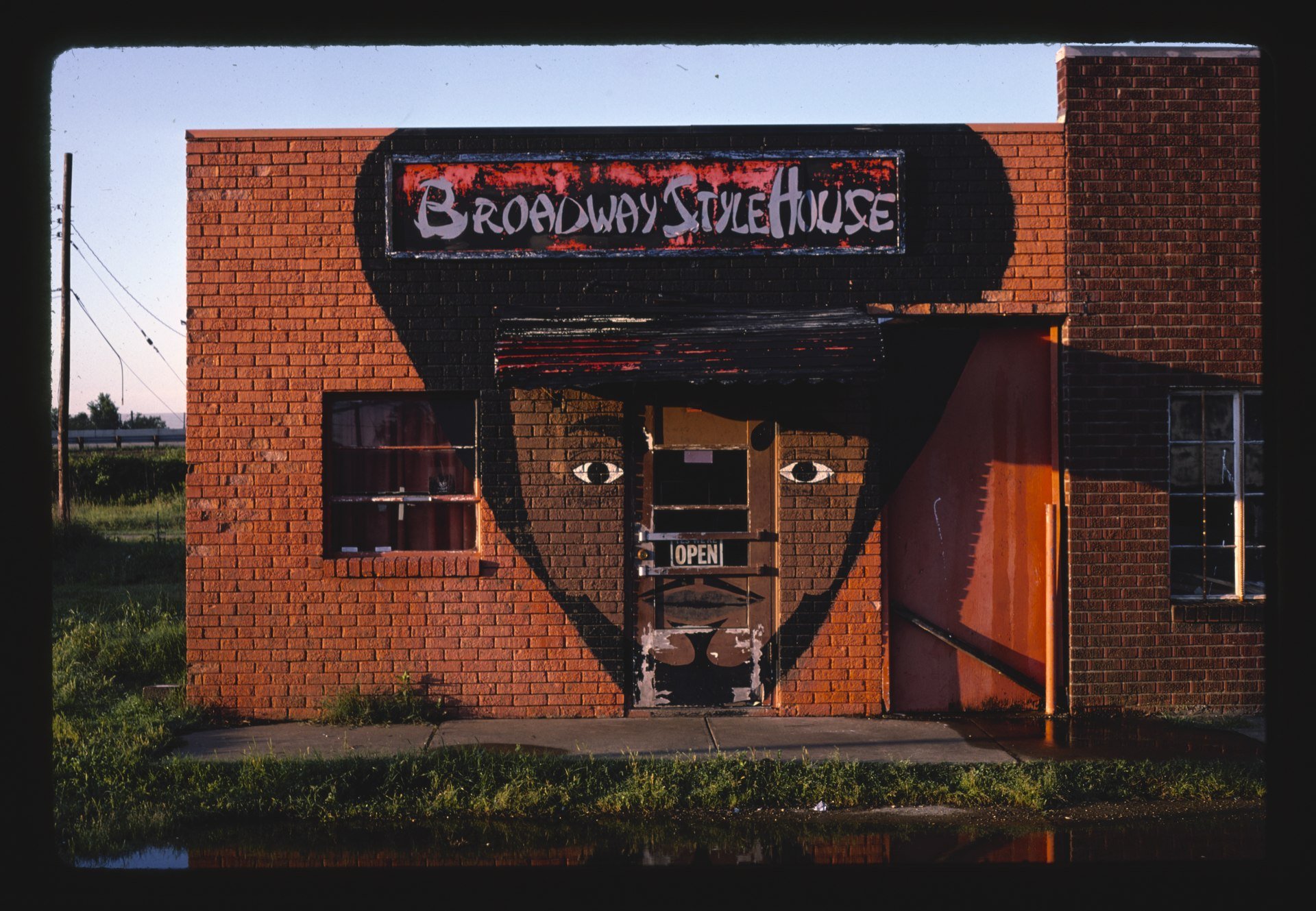 A brick building with a painted face, including eyes, nose, and mouth, on the door. The address sign reads 'Broadway Style House,' and the door has an 'Open' sign. The building features a mural of a person's face painted on the lower part of the wall.