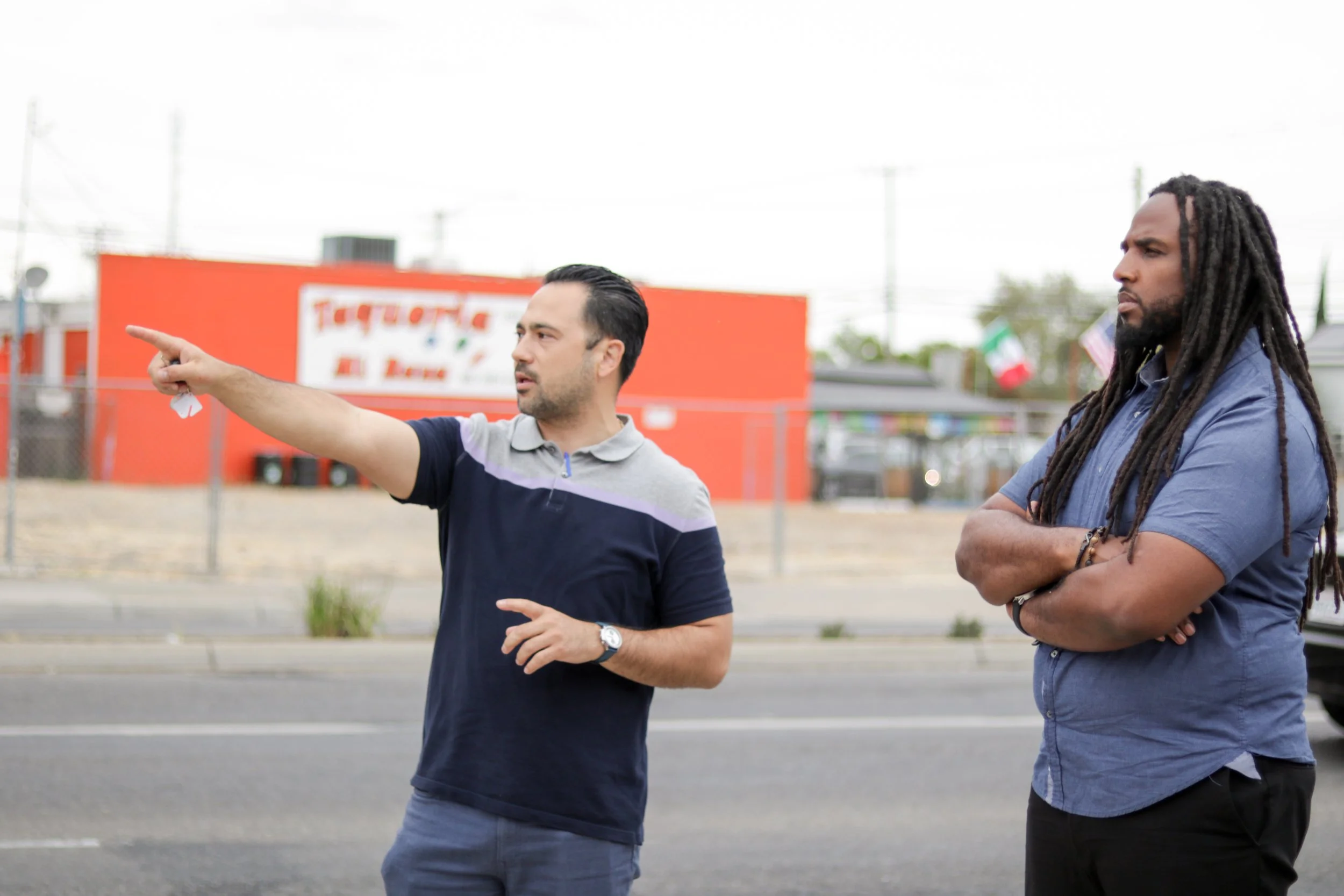 Two men are outdoors having a conversation. One man, who has dark hair and is wearing a polo shirt, is gesturing with his right hand. The other man has long dreadlocks and is wearing a blue shirt with his arms crossed, listening attentively.