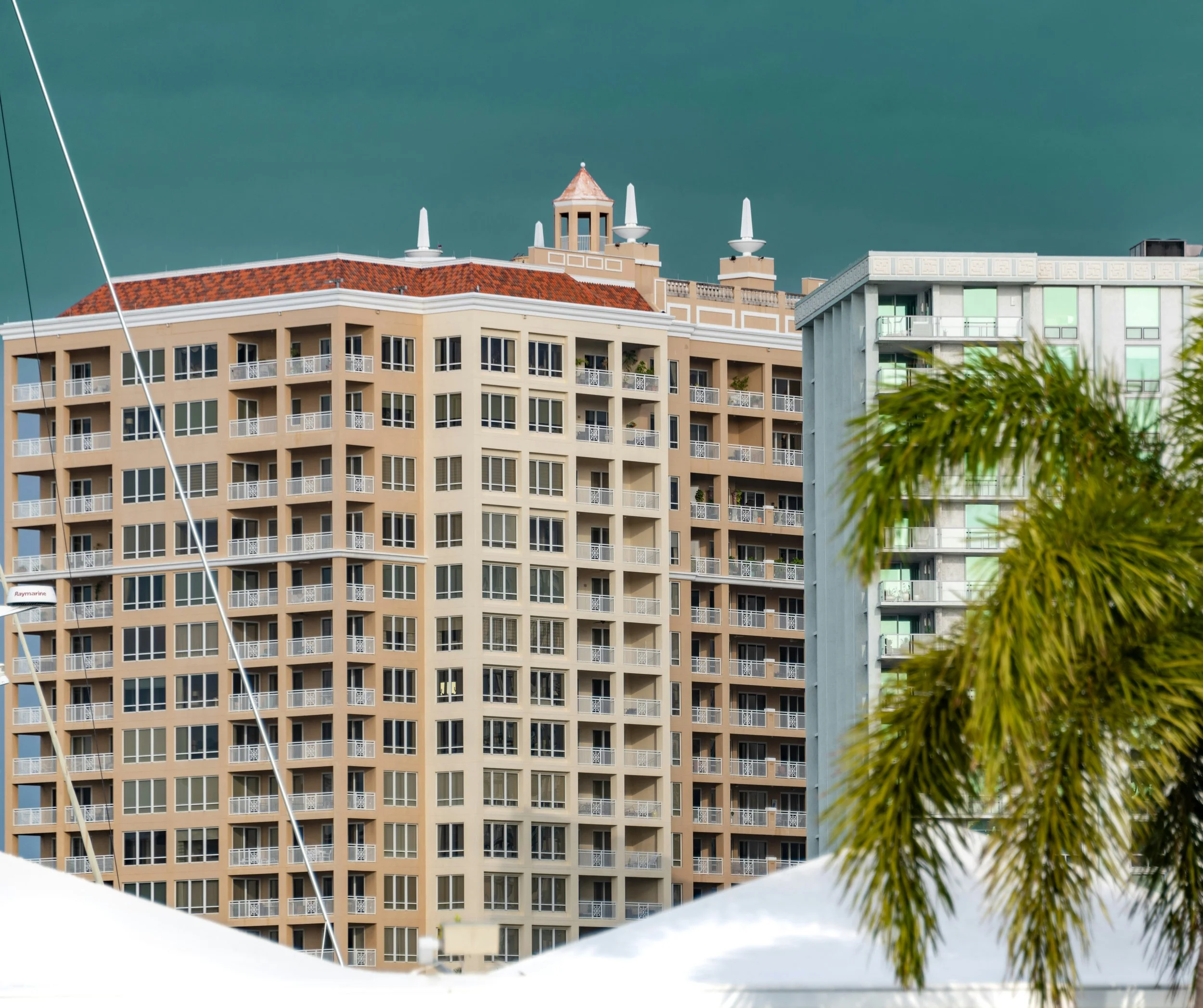 View of a cityscape with multiple high-rise buildings, one with balconies, and a palm tree in the foreground under a cloudy sky.