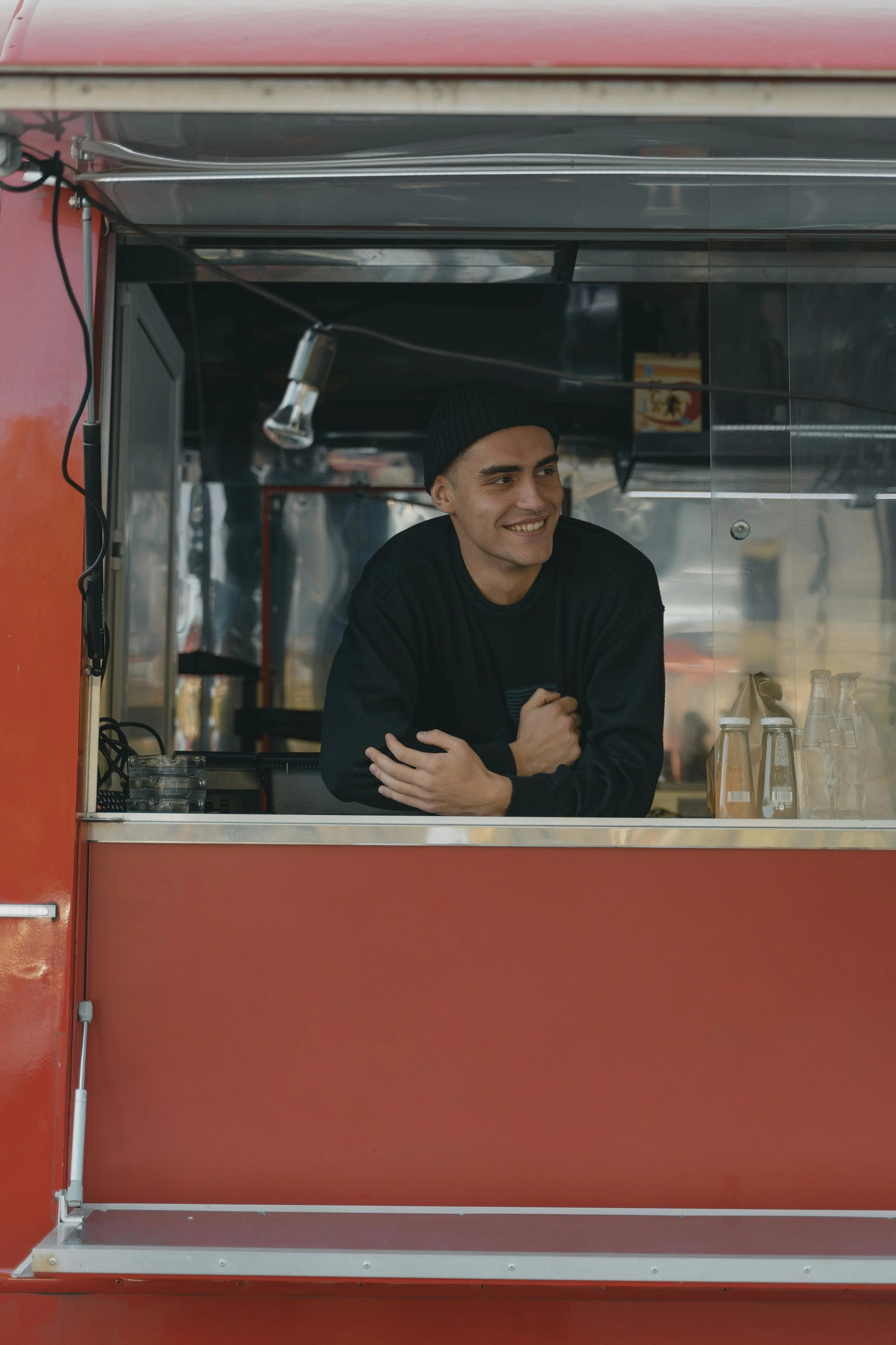 Young man with dark hair, smile, wearing a black beanie and black jacket, leaning on a food truck window, with soda bottles visible on the counter.