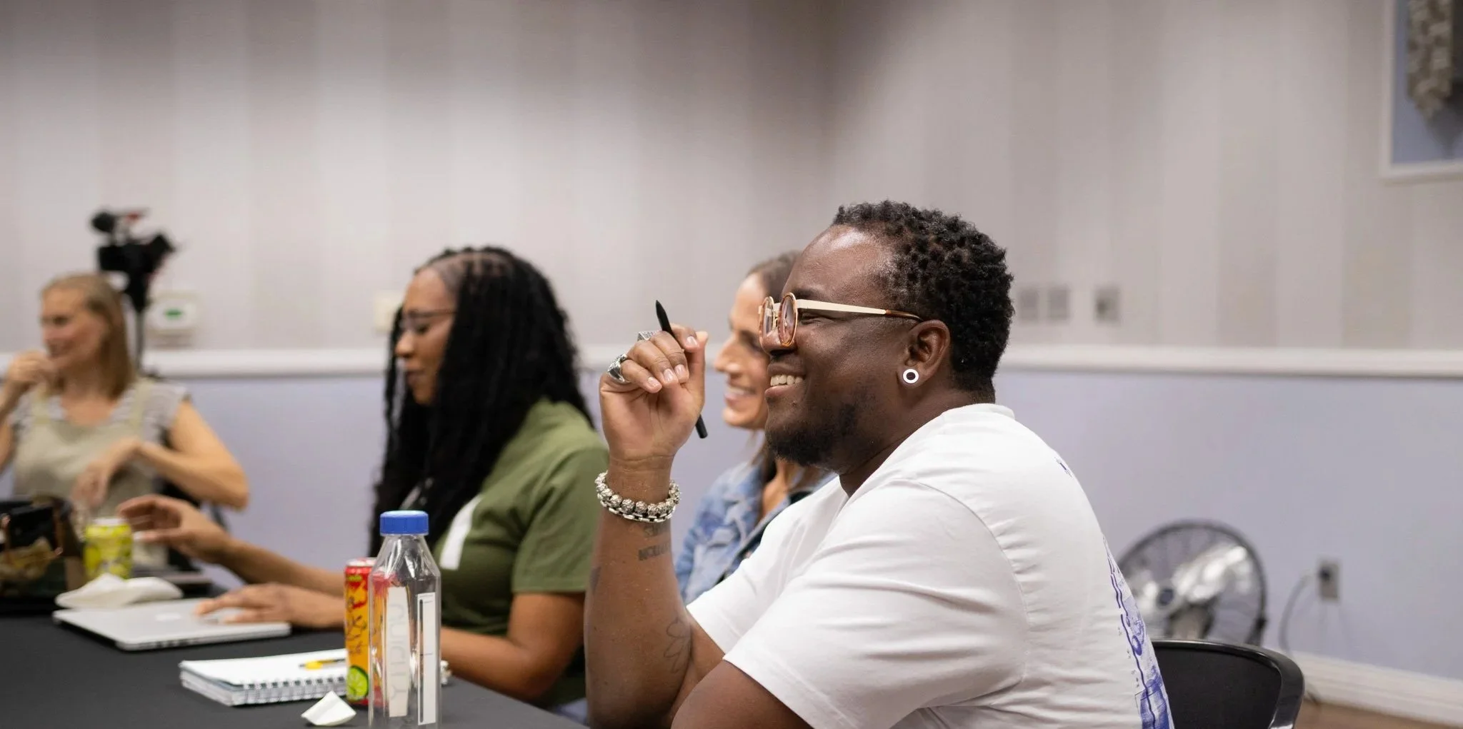 People sitting at a table during a conference or meeting, smiling and engaging, with notebooks, drinks, and a water bottle in front.
