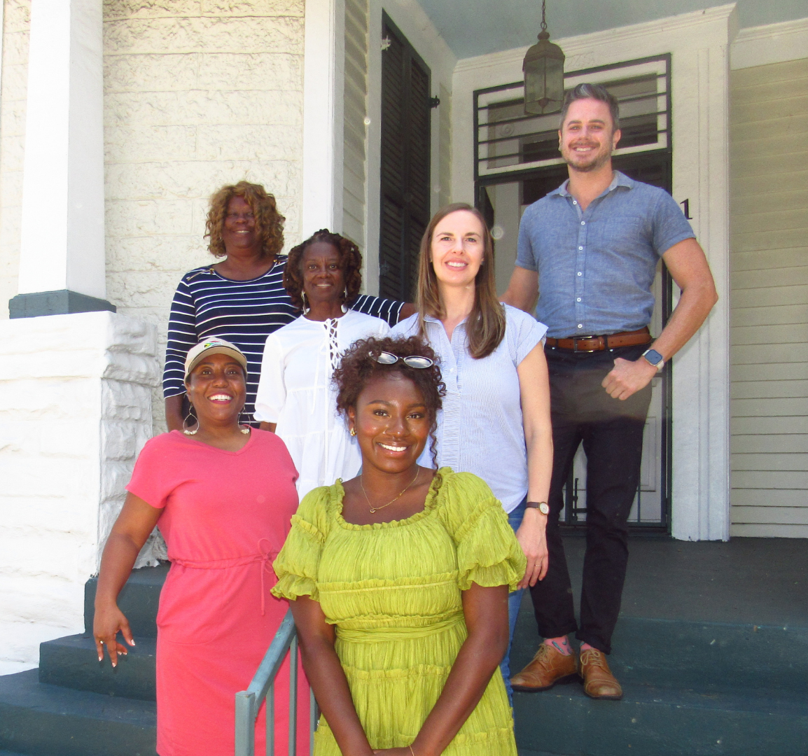 A diverse group of seven women and one man standing on the porch of a house, smiling for the camera.