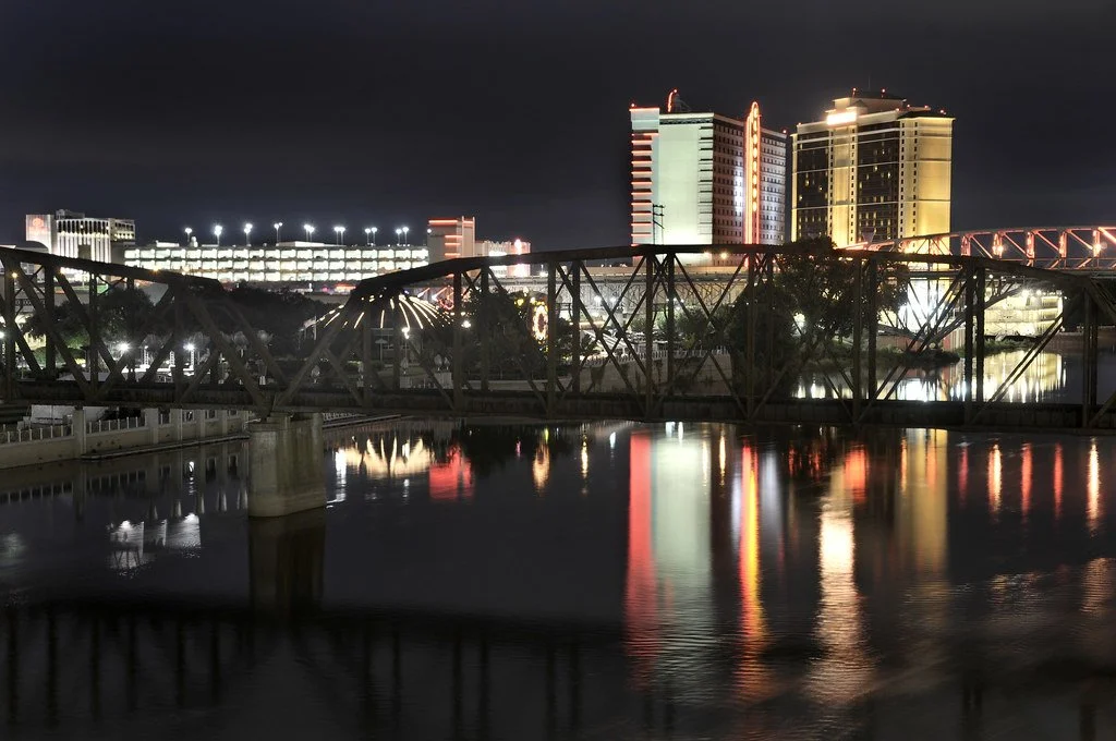Nighttime cityscape with illuminated buildings and their reflections on a river, a bridge in foreground, and a stadium with lights in the background.