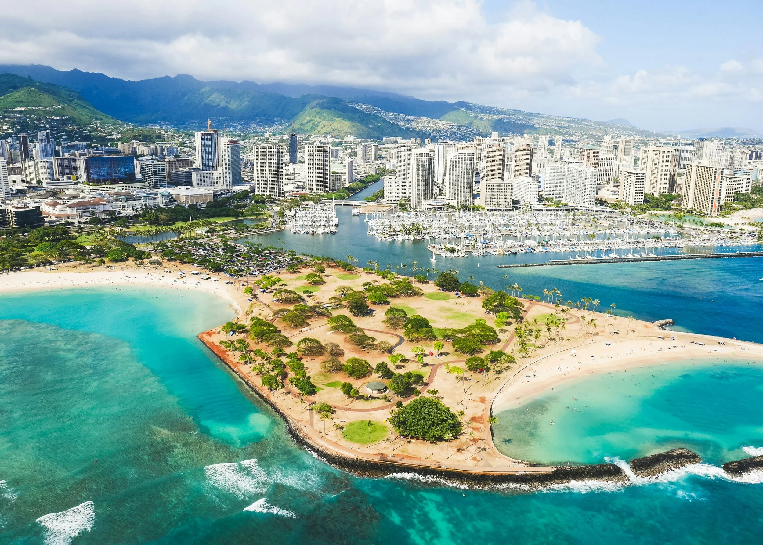 An aerial view of a city with tall buildings, a harbor with boats, a beach with trees, and green mountains in the background.