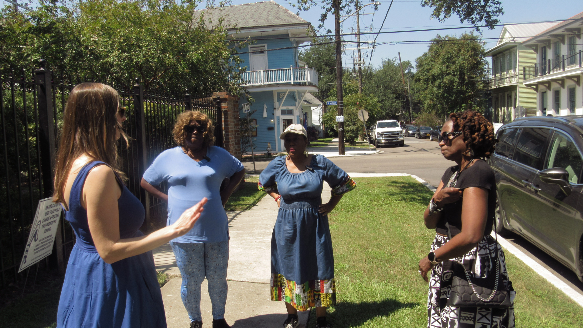 Five women standing on a sidewalk in conversation with a background of residential houses, trees, and parked cars on a sunny day.