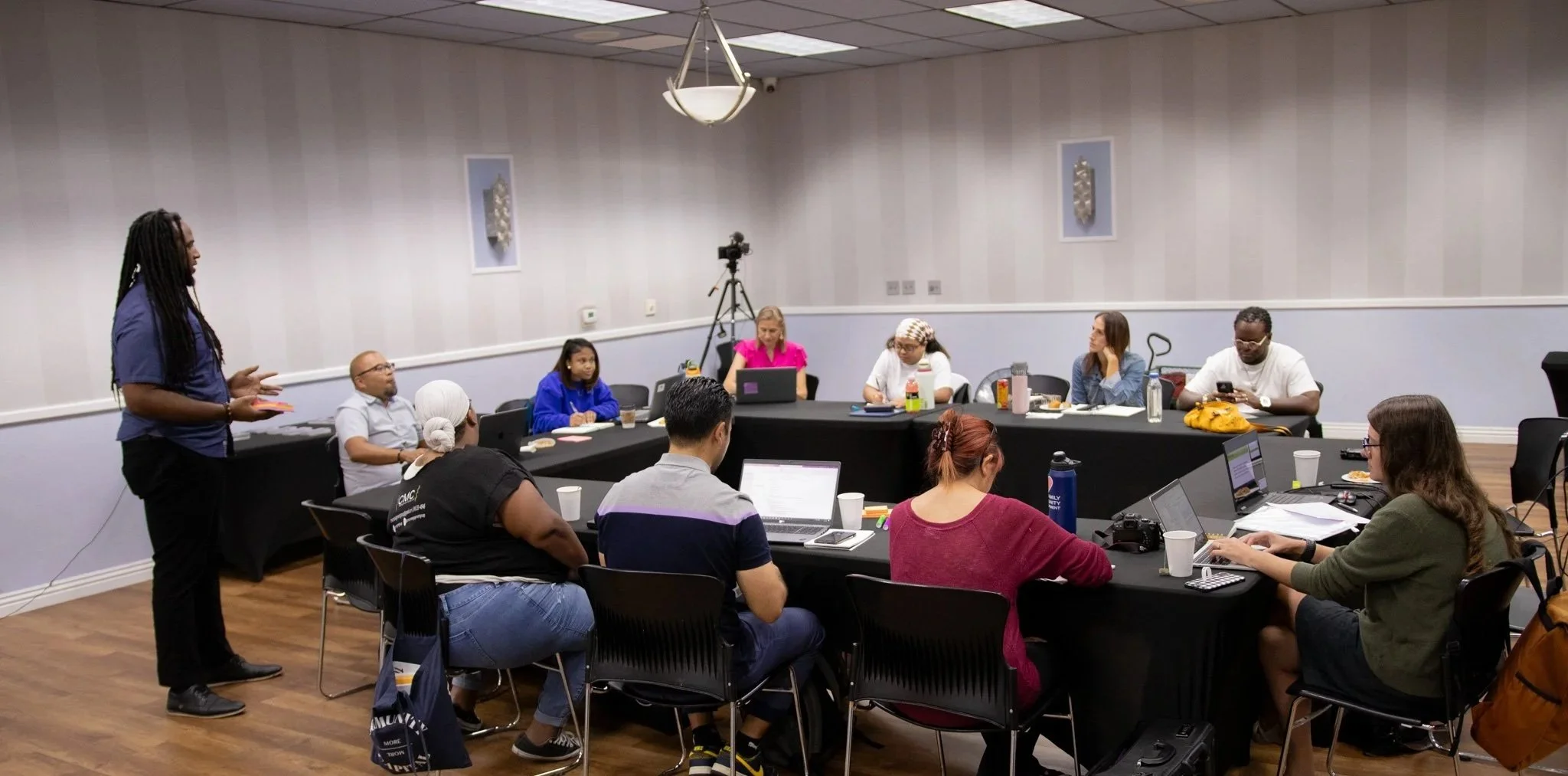 A diverse group of people attending a meeting or workshop in a conference room. One woman is standing and speaking, while others are seated at a large U-shaped table with laptops, notebooks, and drinks.
