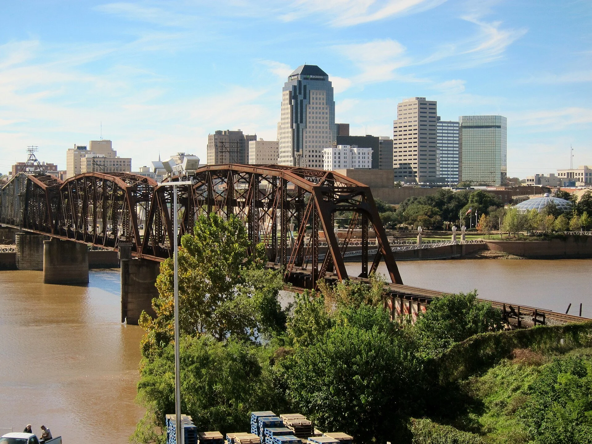 City skyline with tall buildings, a river in the foreground, and a rusted steel bridge crossing the river.