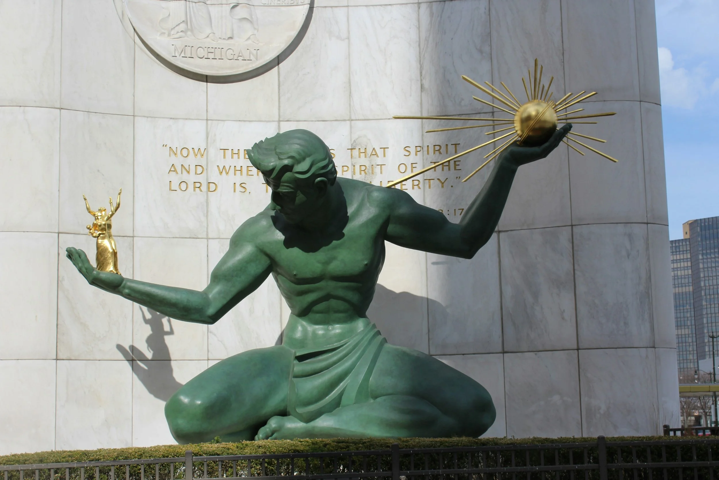 A bronze statue of a muscular man kneeling on one knee with his head bowed, holding a glowing golden globe with rays radiating outward in one hand and a smaller statue of a figure with raised arms in the other hand. The statue is set against a marble wall with engraved text and a Michigan state seal, with a city skyline in the background.