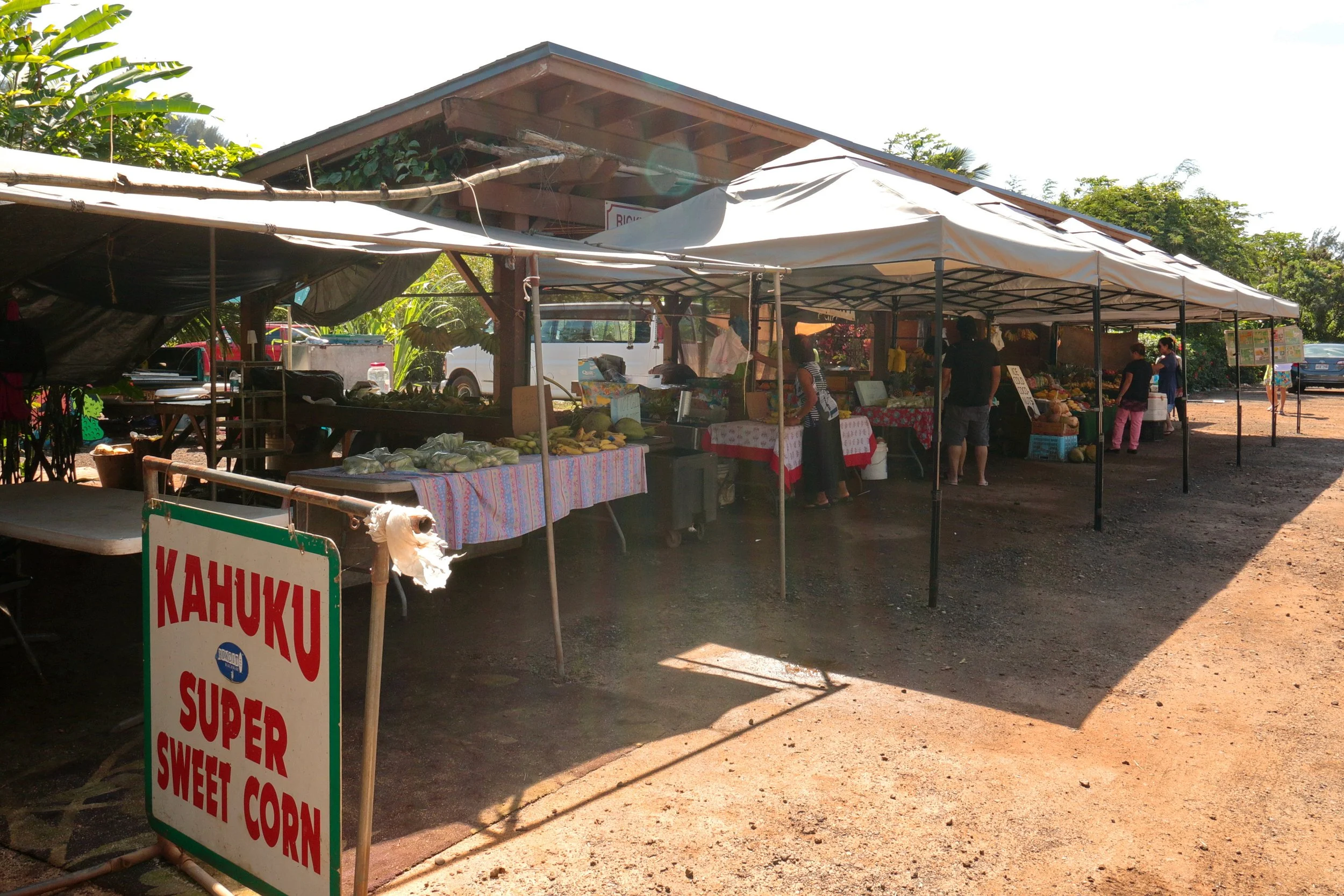 Open-air market with white tents selling produce, people shopping, surrounded by greenery and trees, with cars parked in the background.