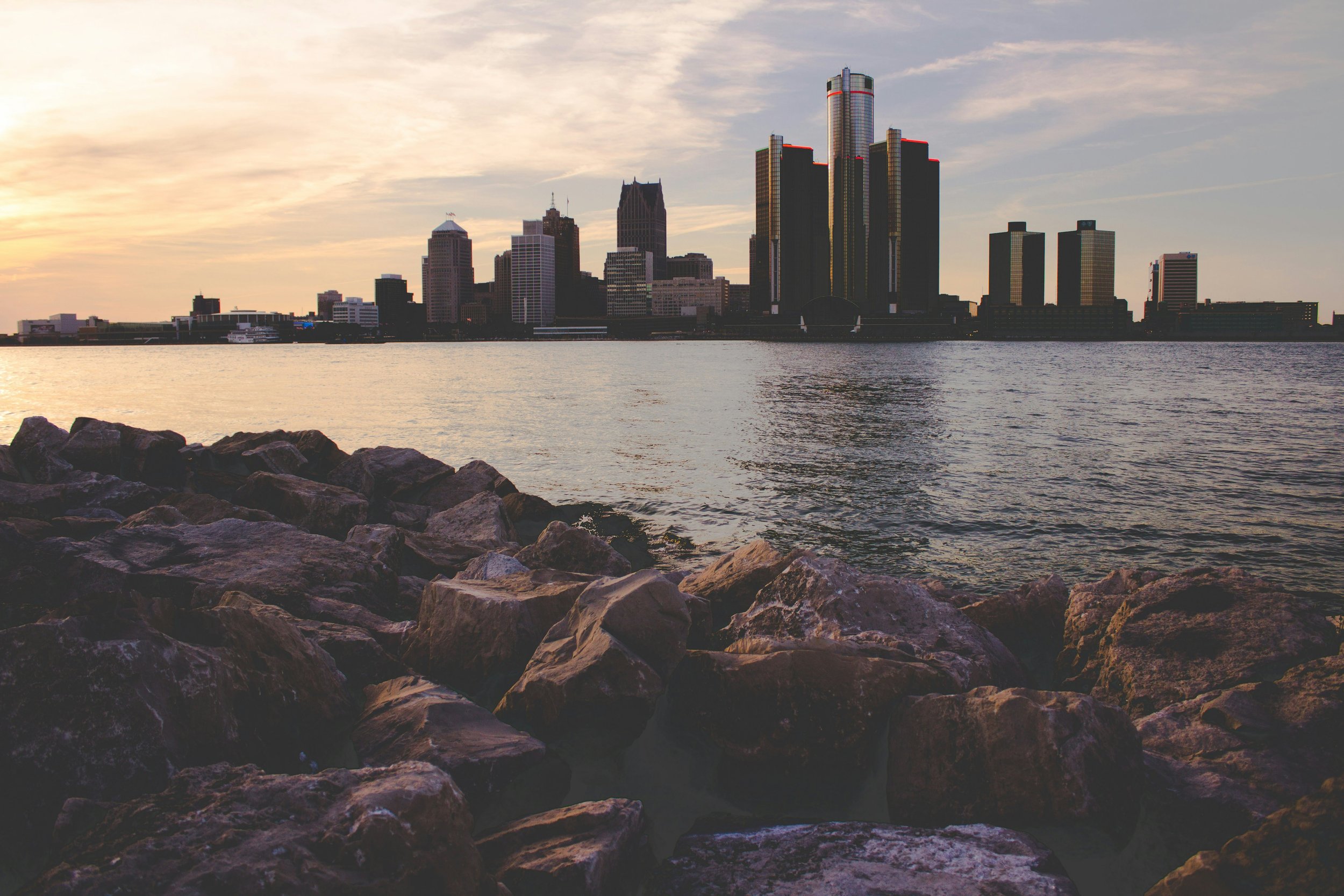 City skyline with tall buildings across a body of water, rocks in the foreground, and a partly cloudy sky at sunset.